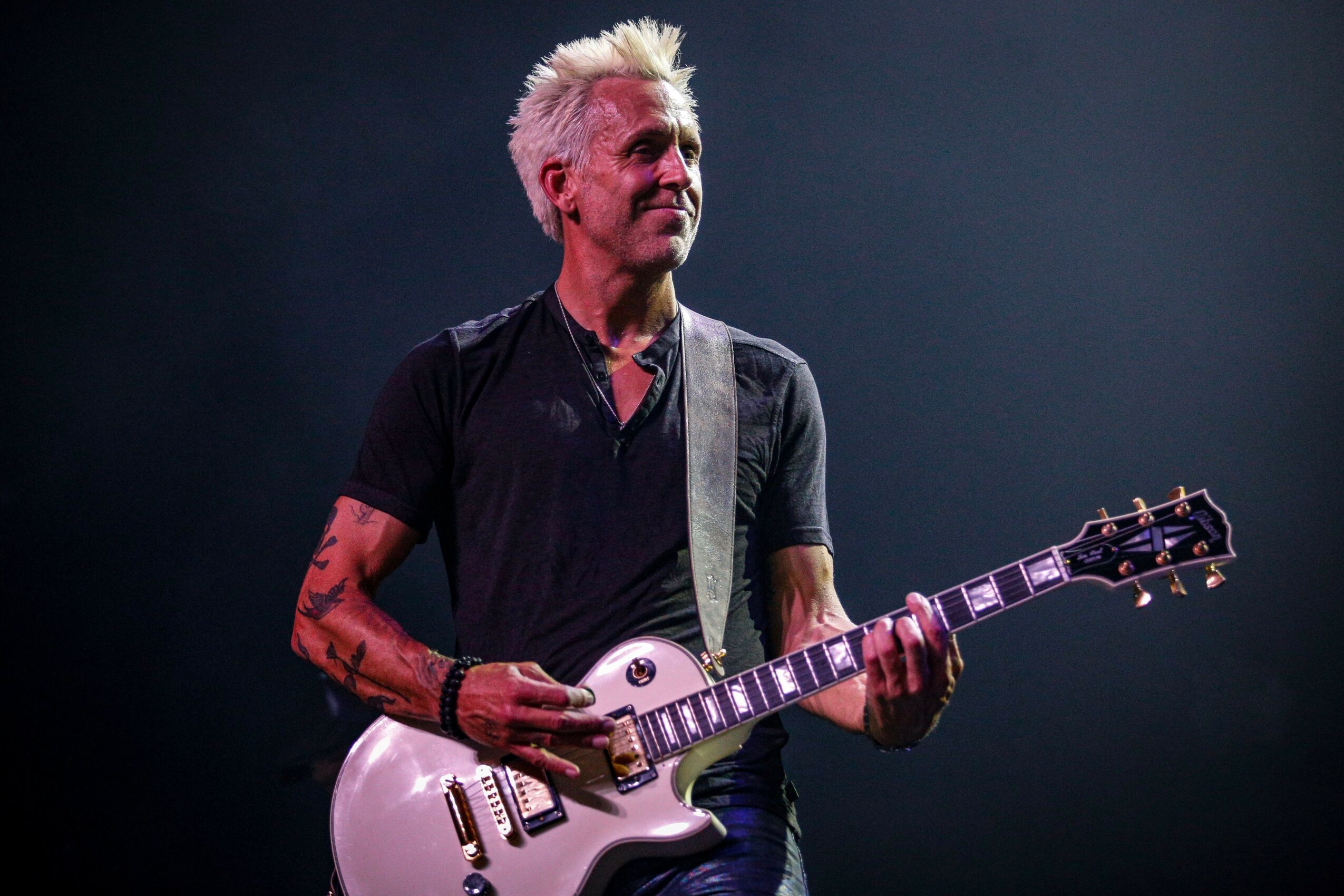 Person with short, spiky blonde hair playing an electric guitar on stage, wearing a black shirt and tattoos visible on arms.