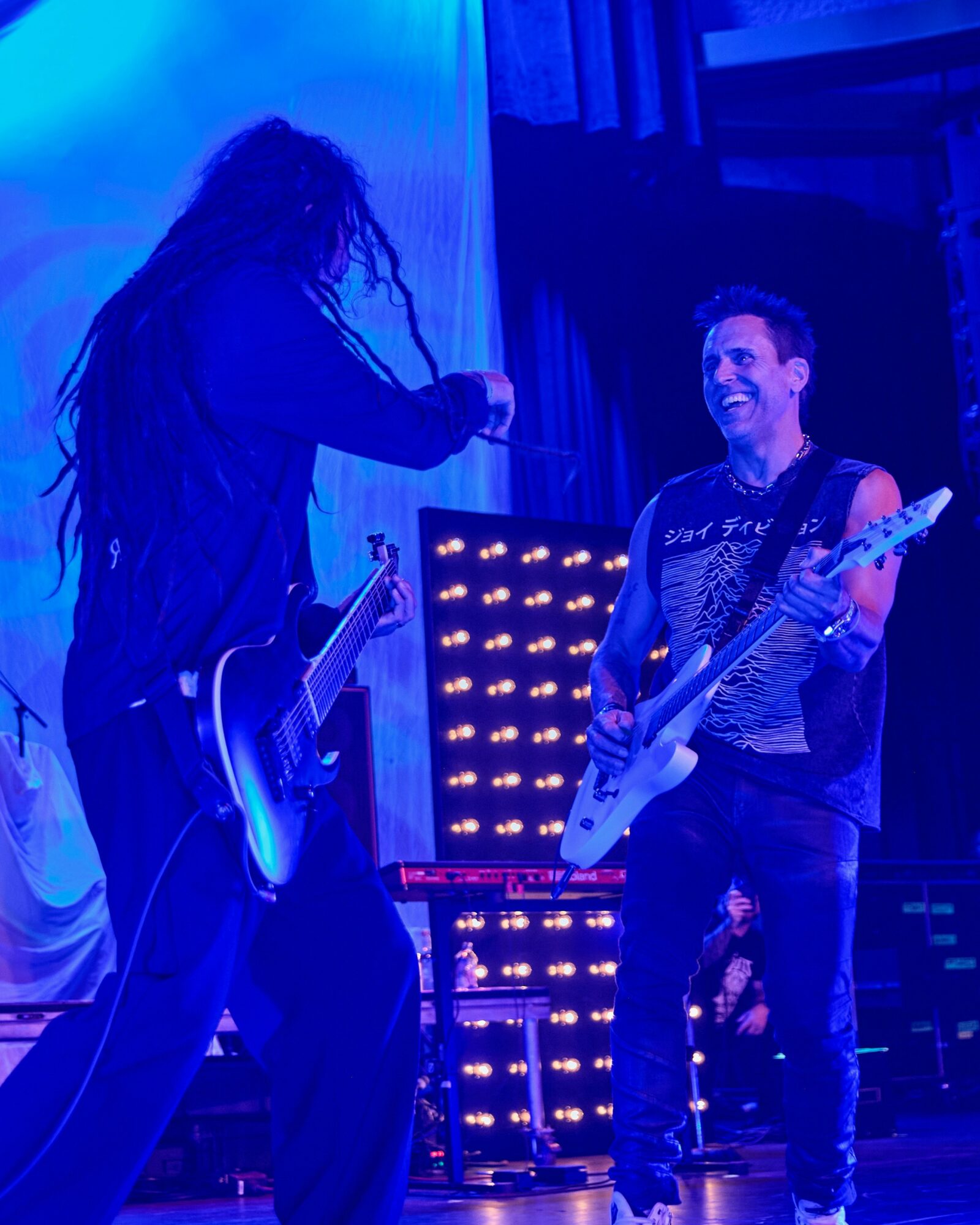 Two musicians on stage, one with long dreadlocks and the other smiling, both holding electric guitars, with stage lights behind them.