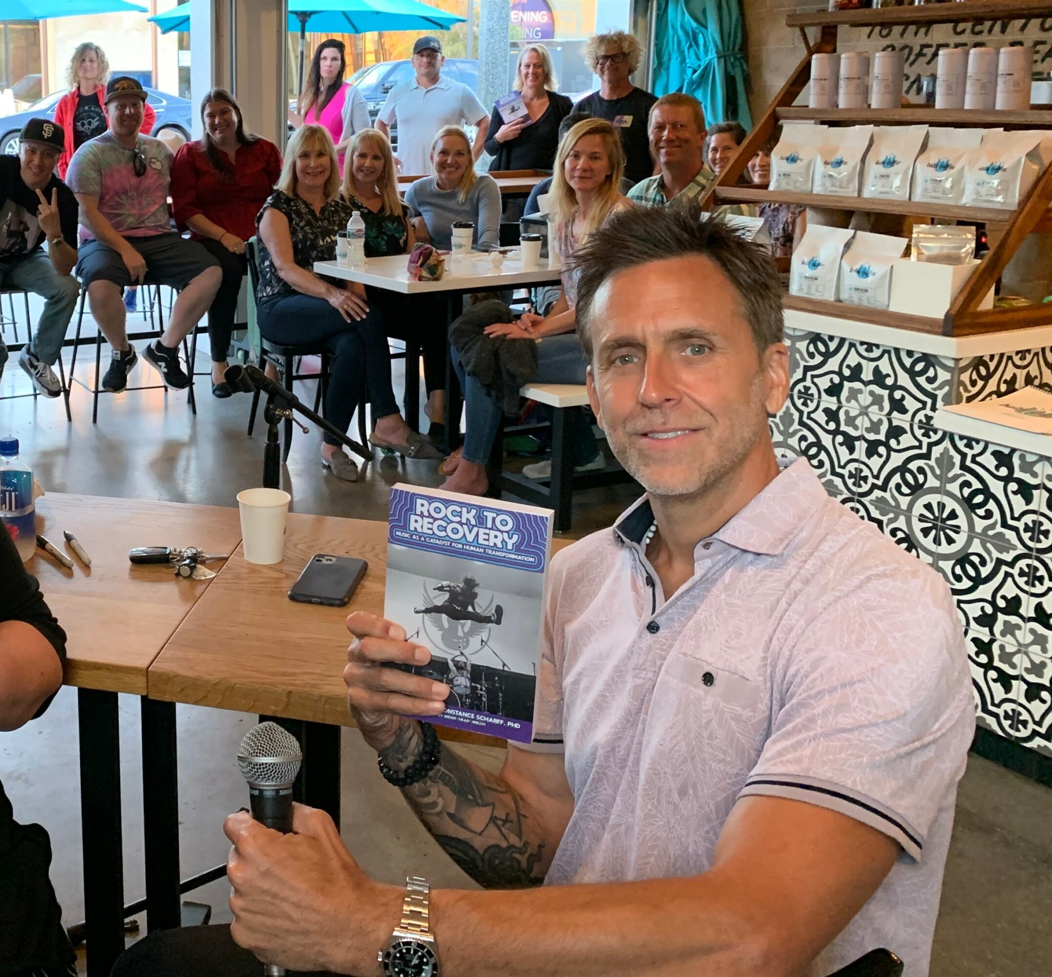 Man holding a book titled 'Rock to Recovery' in a cafe with a group of people in the background.