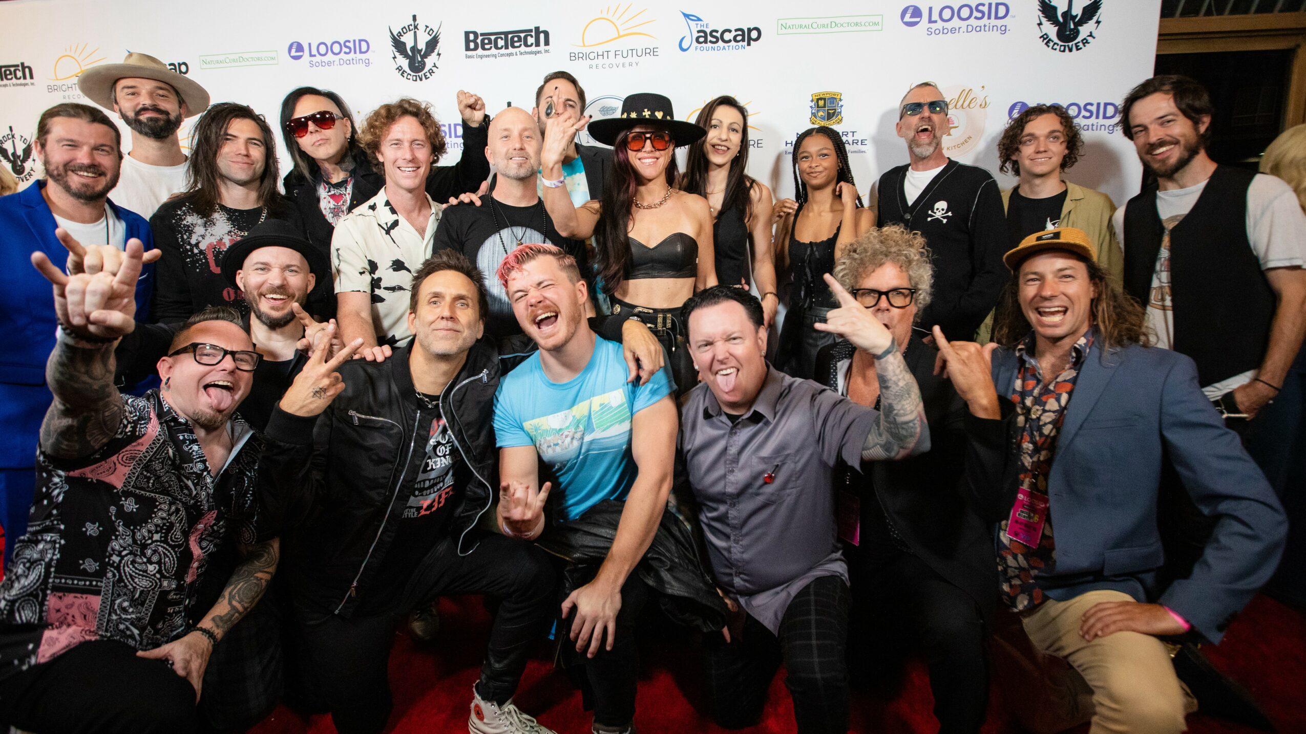 Group of people posing on red carpet, smiling, some making hand gestures, with event backdrop behind them.
