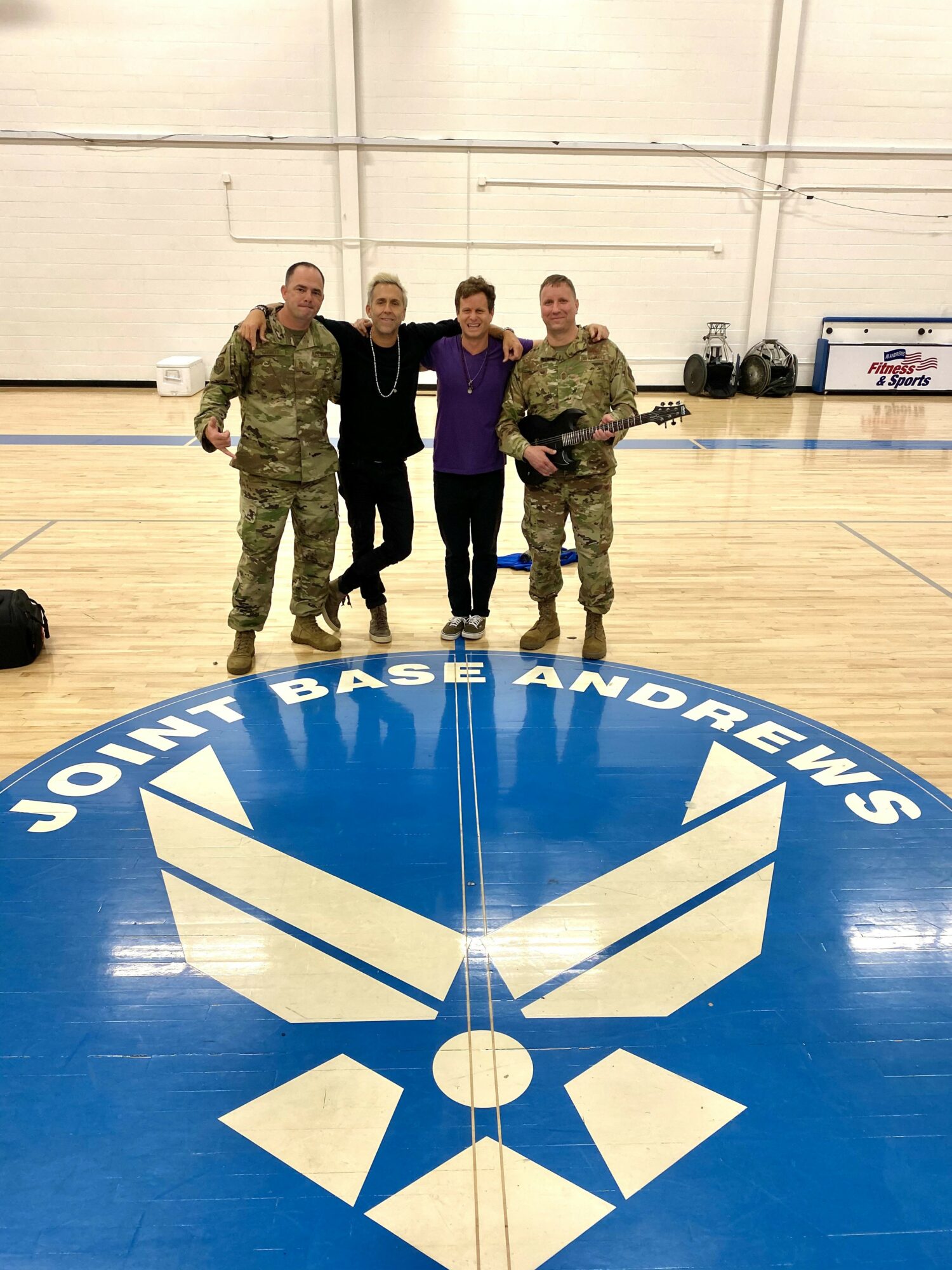 Four people standing on a gym floor near a large circular logo with text, two in military uniforms and two in civilian clothing.
