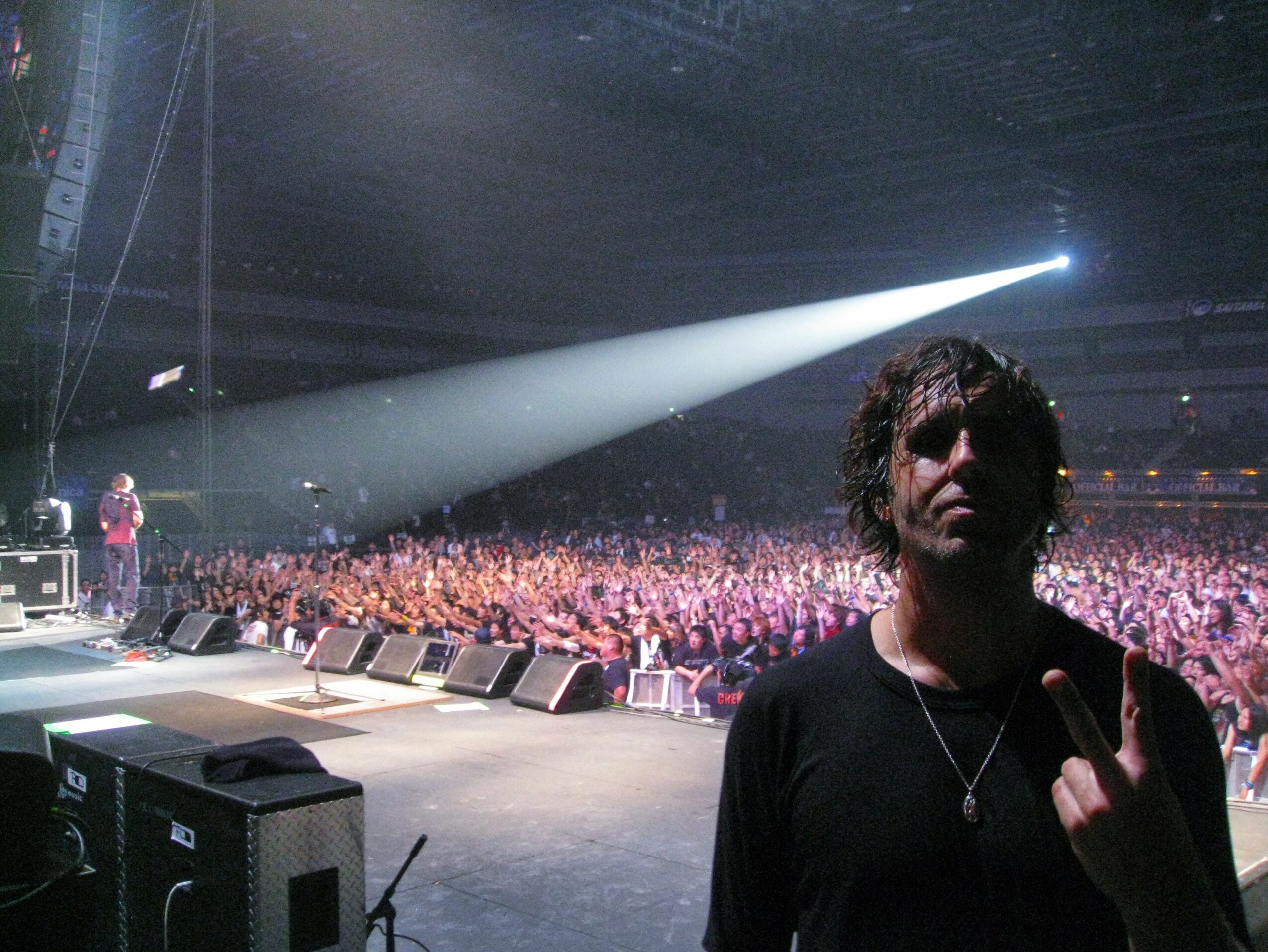 Person with dark hair and black shirt in front of a large crowd at a concert, holding up a hand with middle finger raised, stage lights shining behind.
