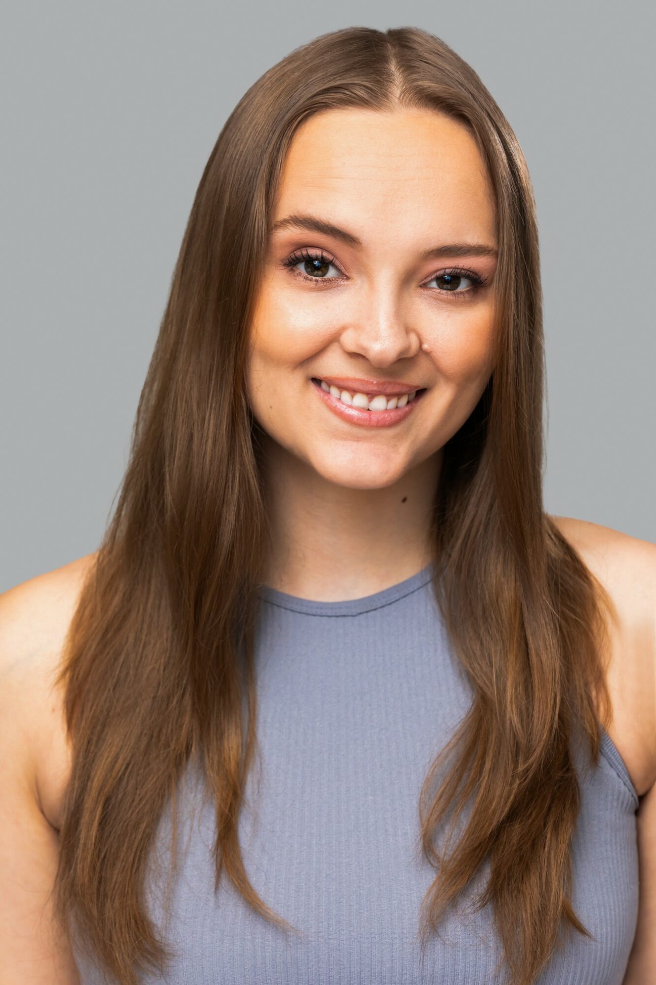 Young woman with long brown hair smiling, wearing a sleeveless blue top, against a gray background.