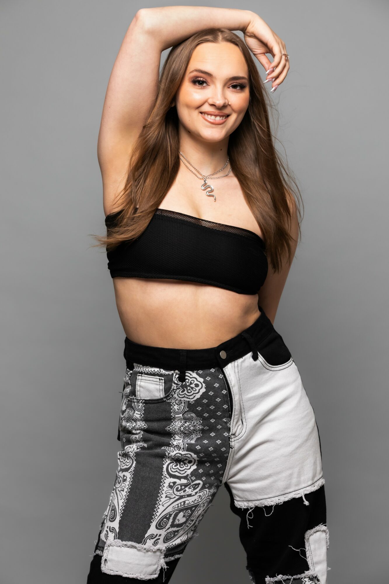 Young woman with long hair smiling, wearing a black crop top and patterned pants, posing with one arm raised.