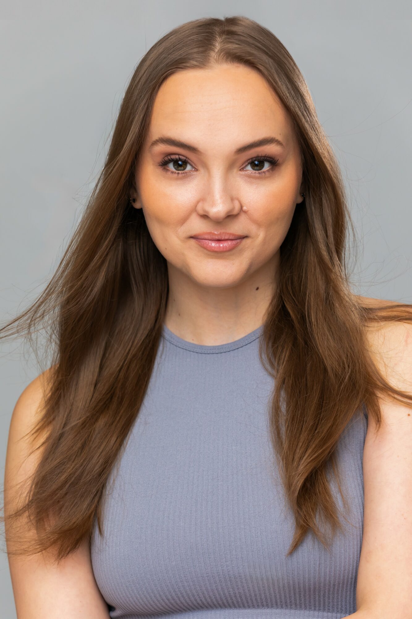 Young woman with long brown hair wearing a sleeveless blue top, smiling slightly, against a light gray background.