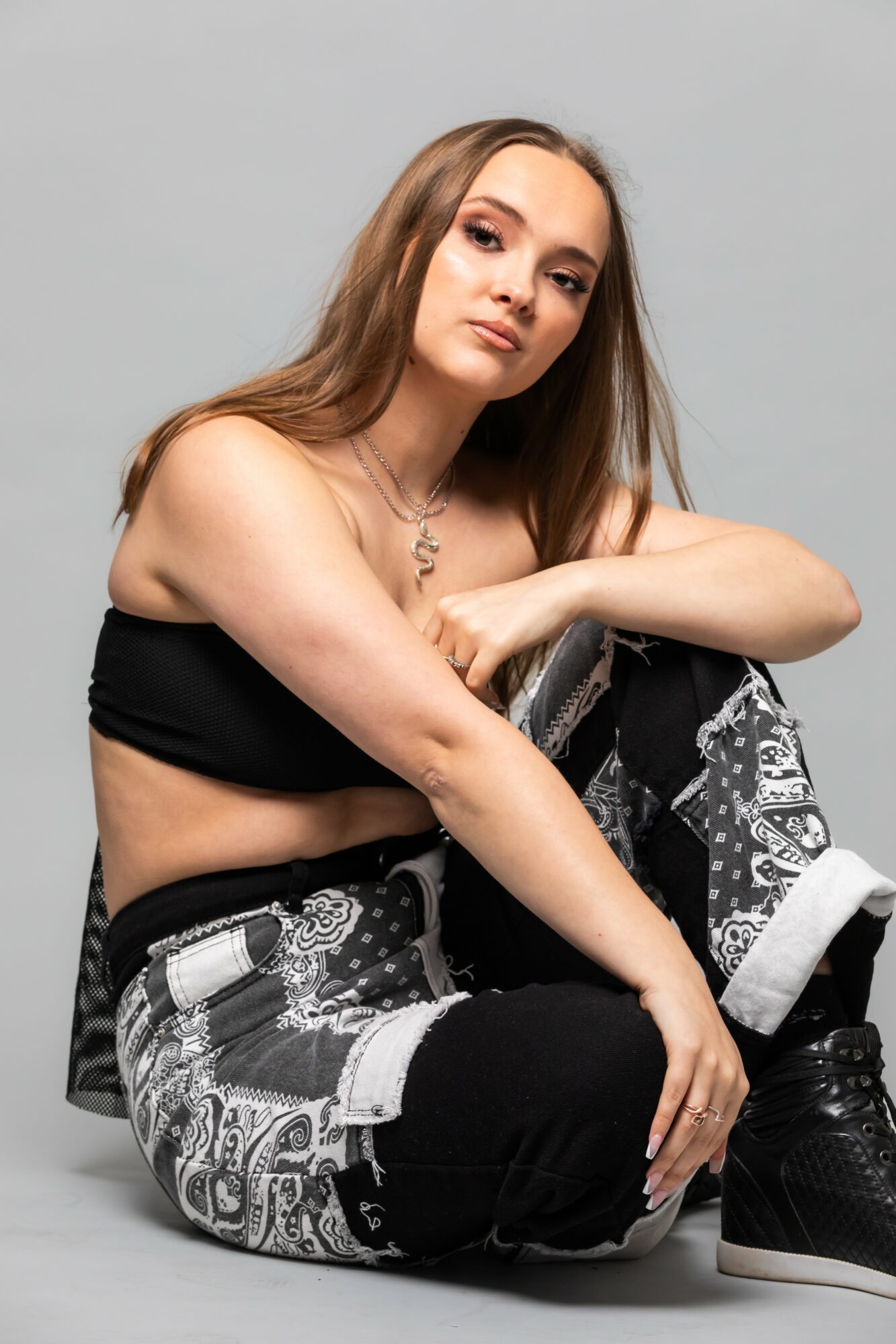 Young woman with long hair sitting on the floor, wearing a black crop top and patterned pants, against a plain background.