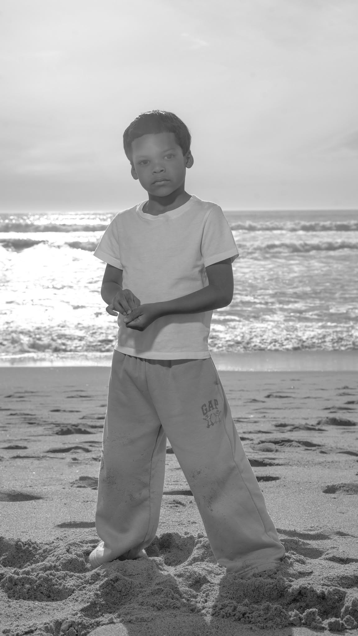 Child standing on beach with ocean waves in background, wearing a t-shirt and loose pants, looking at the camera.