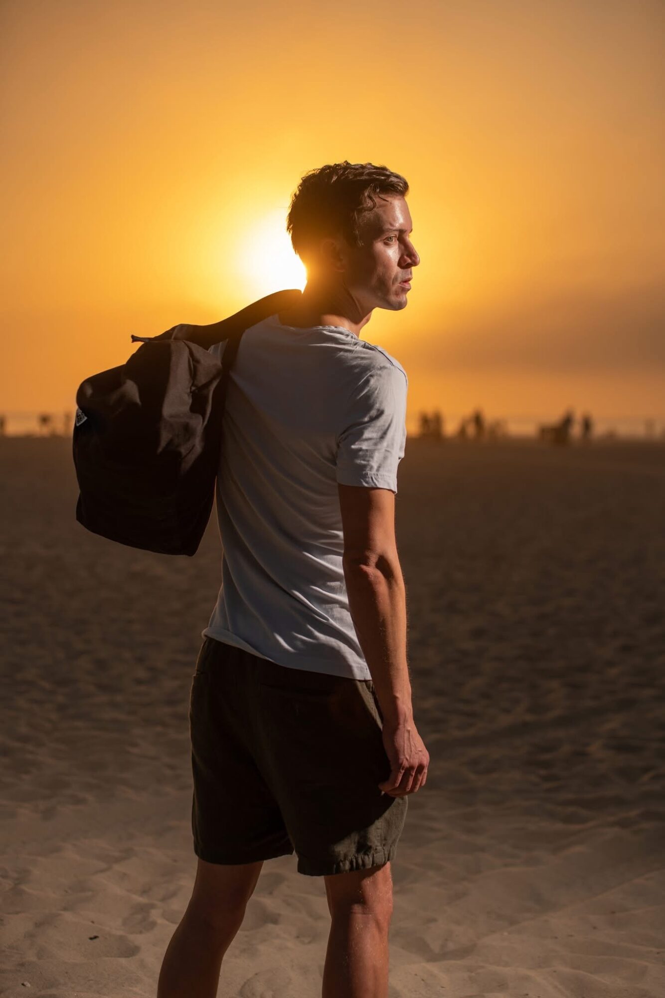 Young man standing on a beach at sunset, carrying a backpack, looking to the side, with a warm orange sky.