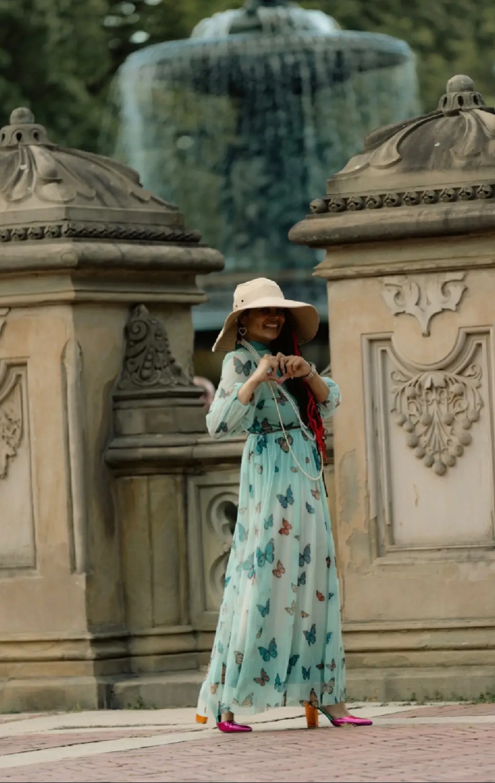 Woman in a long blue dress and wide-brimmed hat stands near ornate stone structures with a fountain in the background.