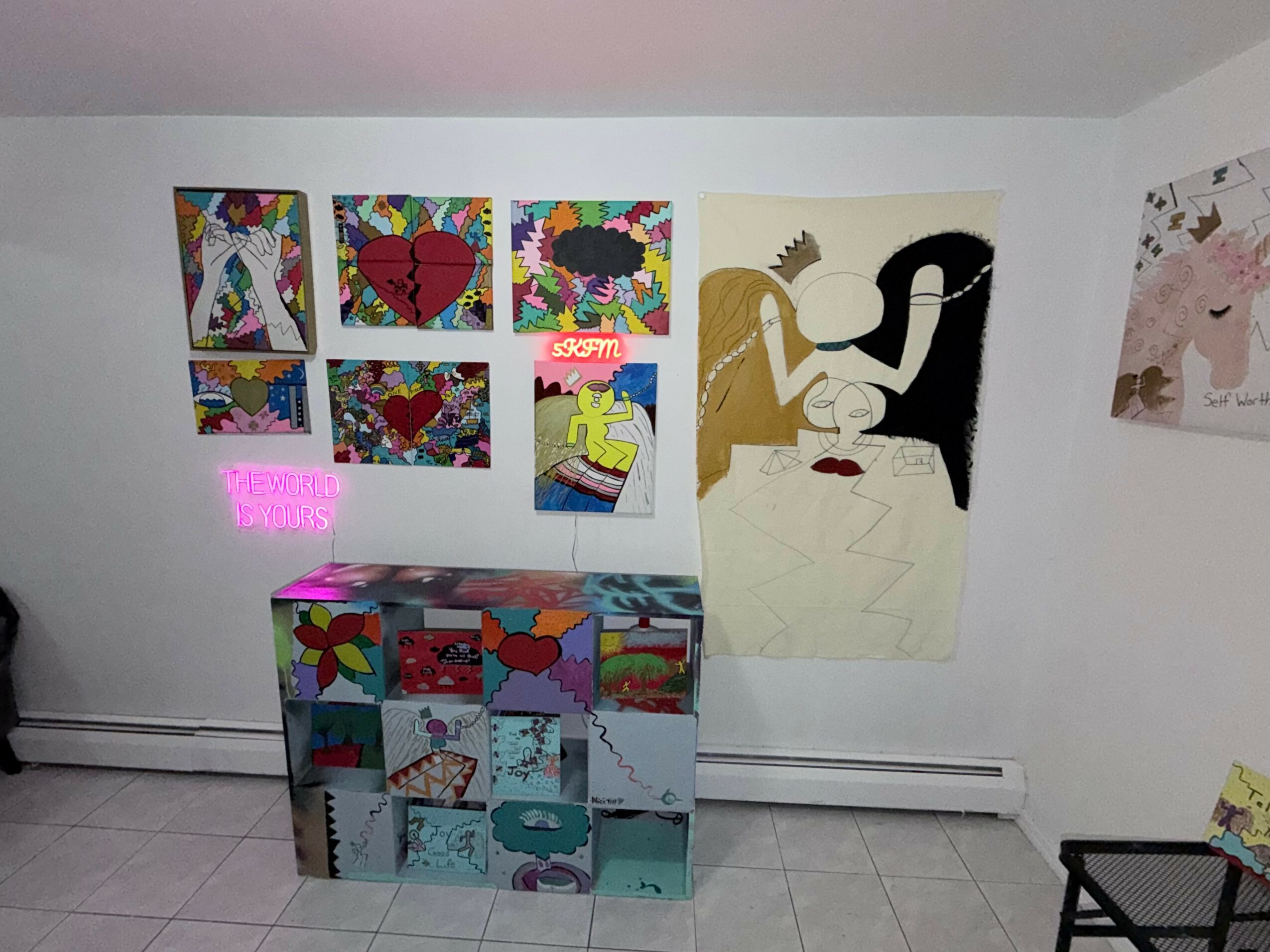 Wall with colorful artwork, a large painting of two abstract figures, and a decorated cabinet below. A small pink sign is visible.