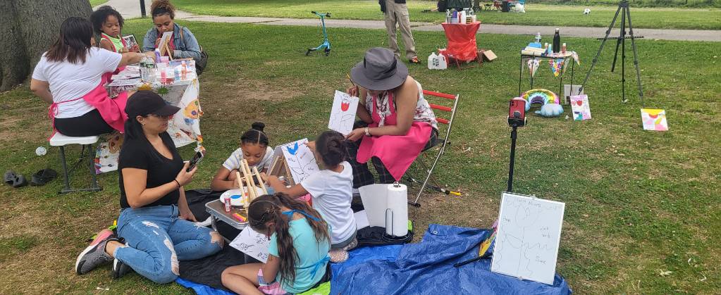 Group of children and adults sitting on grass at outdoor event, some drawing and engaging in activities, with tables and equipment nearby.