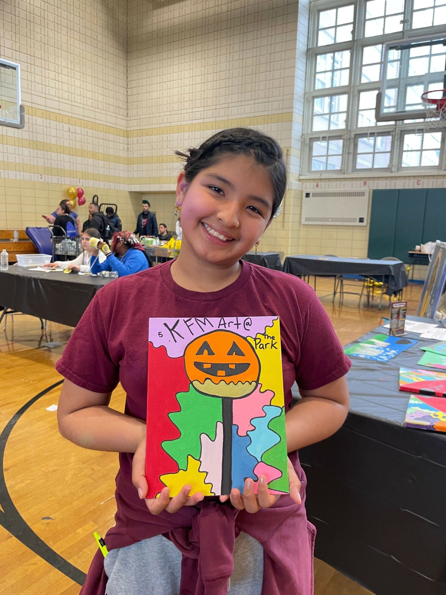 Girl smiling holding a colorful Halloween-themed drawing with a pumpkin, tree, and clouds in a school gymnasium.