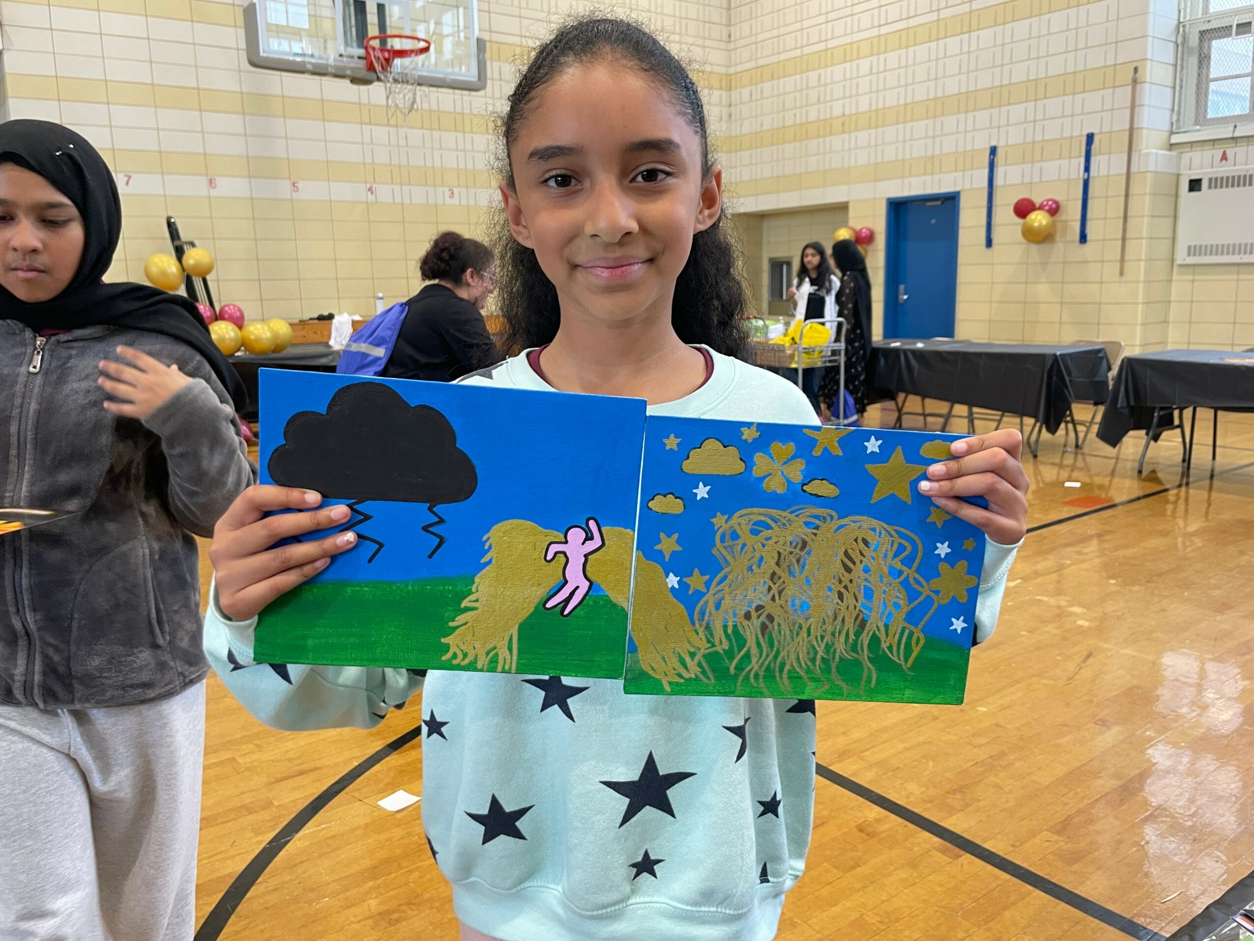 Girl holding a colorful drawing of a pink unicorn, clouds, stars, and a lion in a gymnasium.
