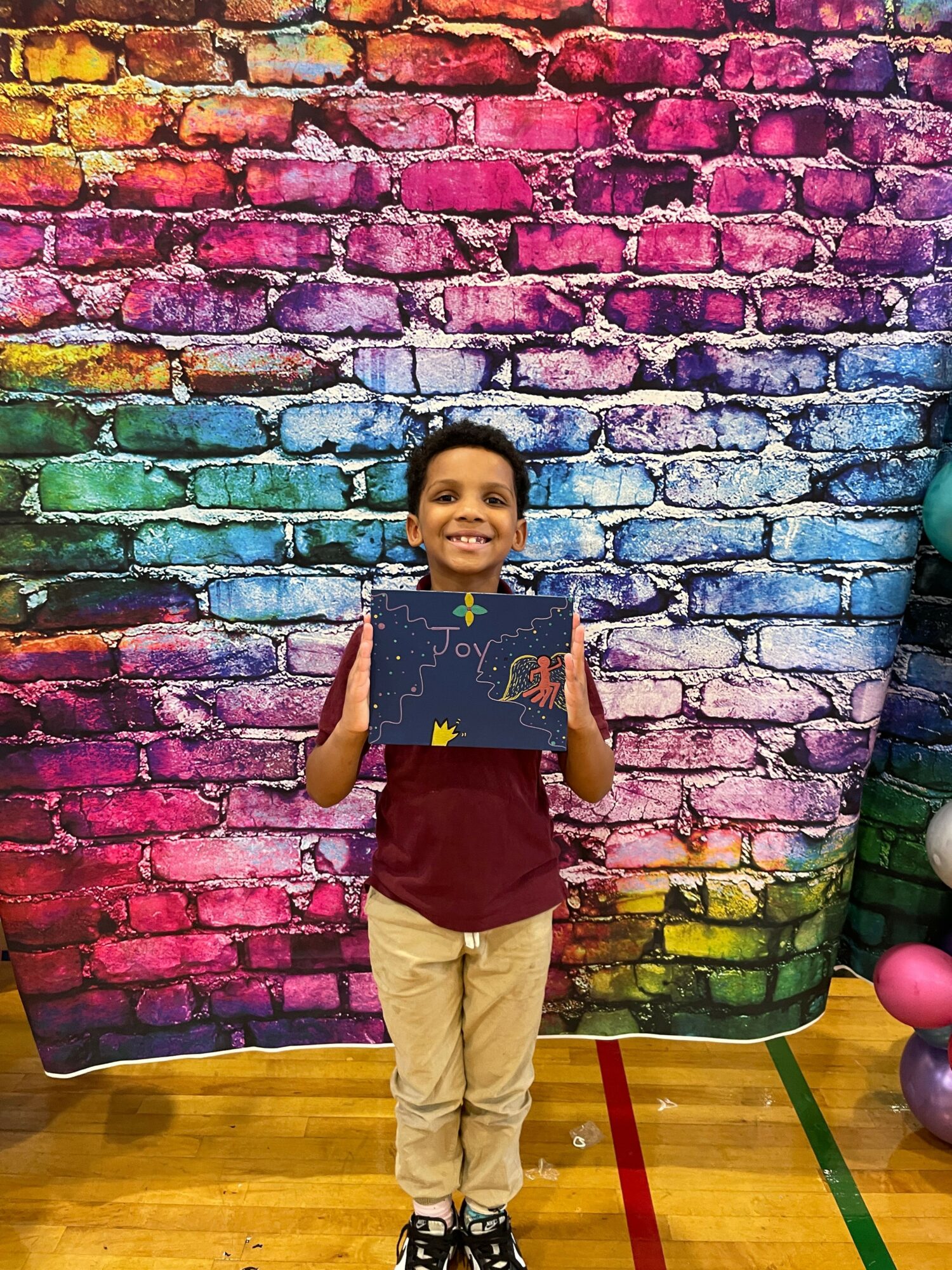 Child standing in front of a colorful brick wall, holding a book, wearing a navy shirt and beige pants.
