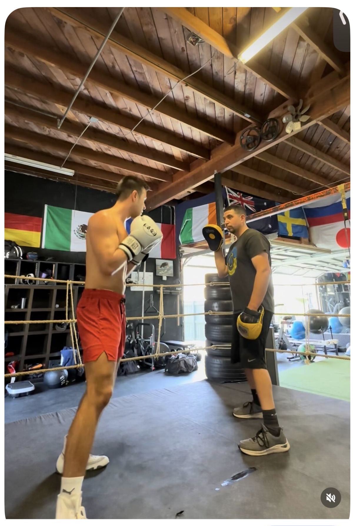 Two men in boxing gloves face each other in a boxing gym with flags hanging from the ceiling.