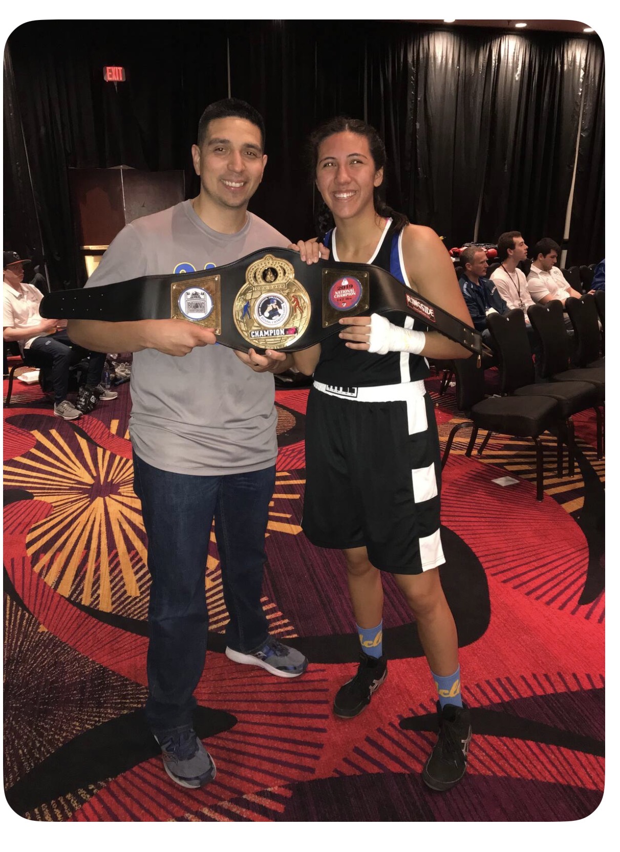 Two people holding a championship belt, smiling, in a room with black curtains and seated audience, colorful carpet.