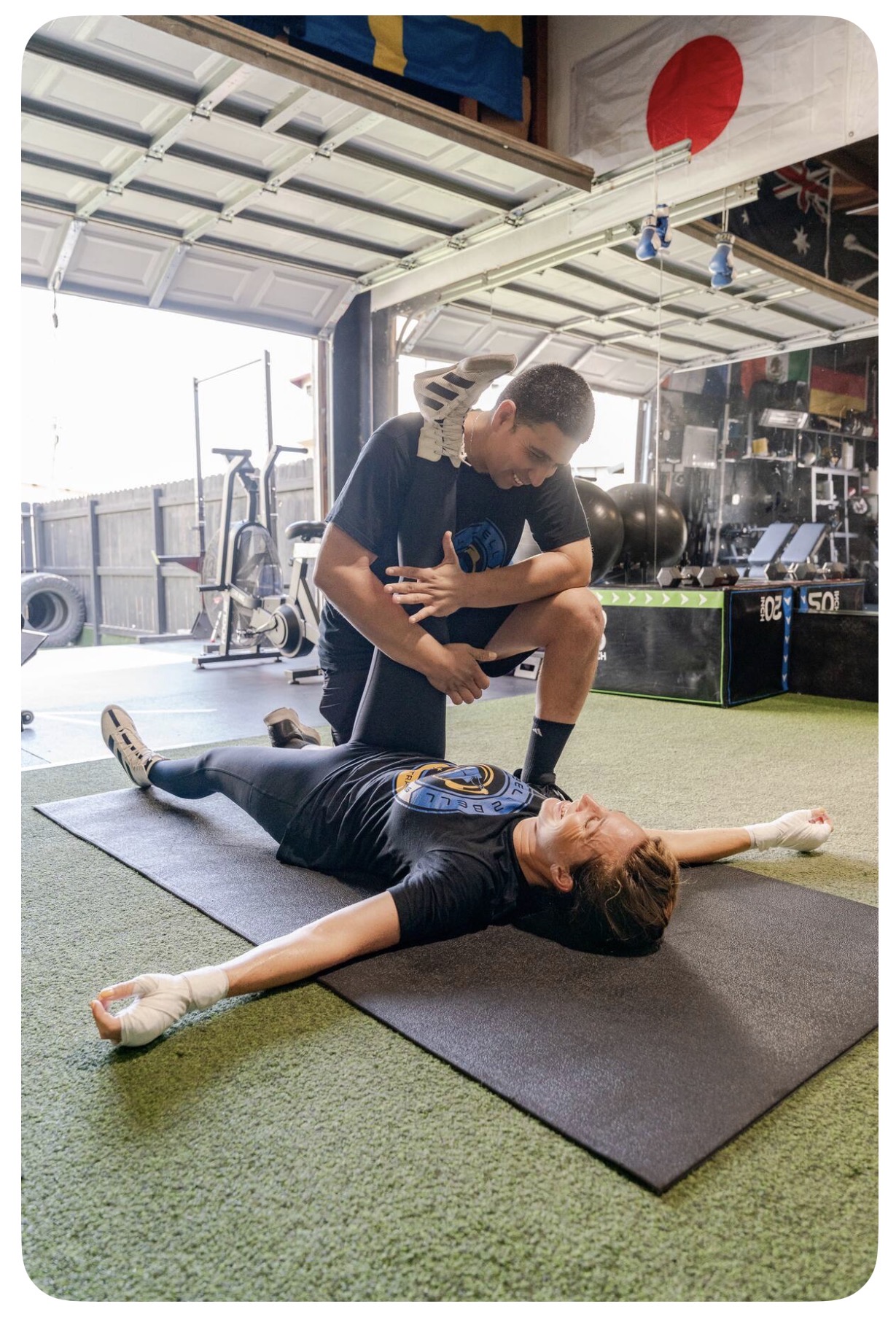 Trainer assisting a woman stretching on a mat in a gym with exercise equipment and Japanese flag in background.