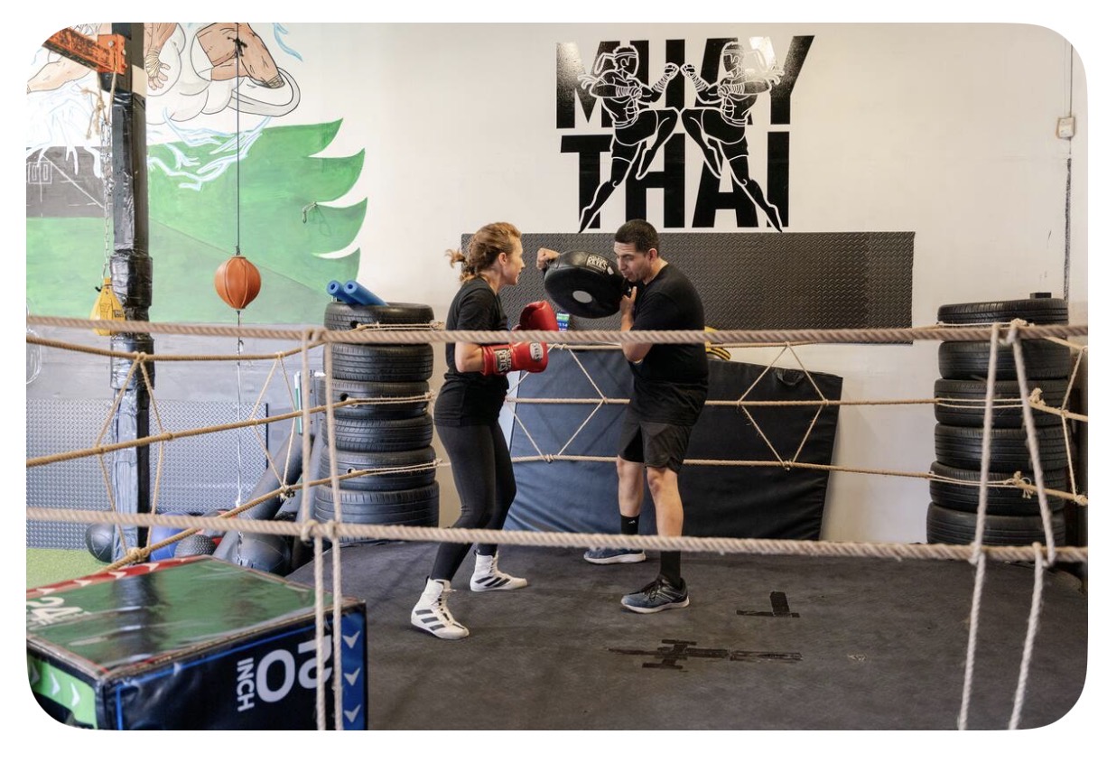 Two people in boxing gloves training in a boxing ring with tires and a wall with graffiti art behind them.