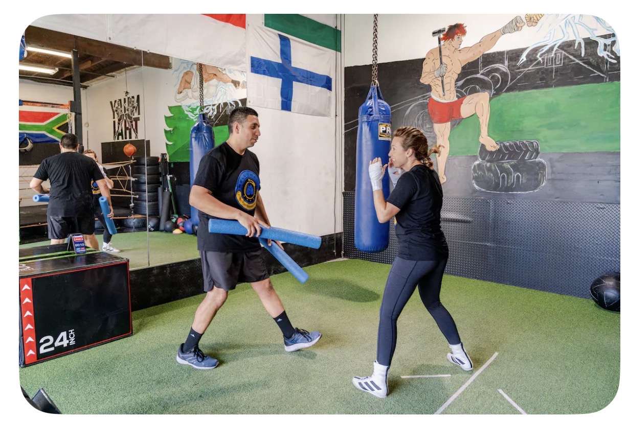 Two people training with a punching bag in a gym, with a mural and flags on the wall behind them.