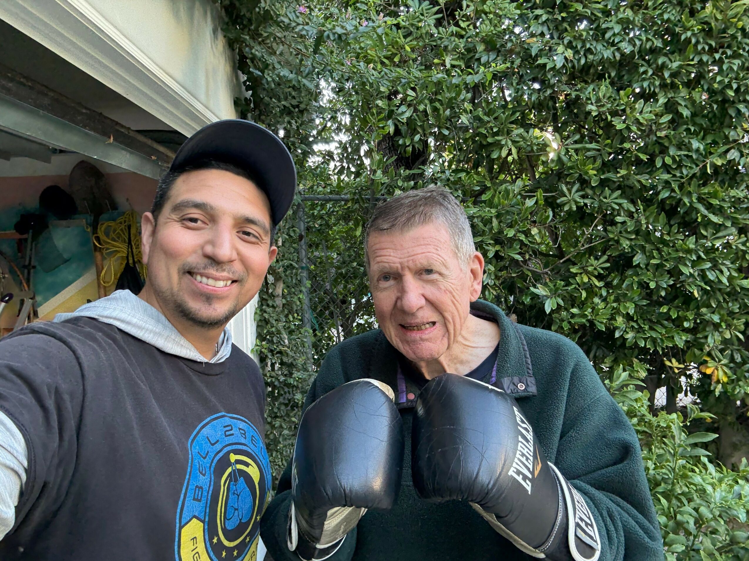 Two people taking a selfie outdoors with greenery in the background, one wearing boxing gloves and the other wearing a cap.