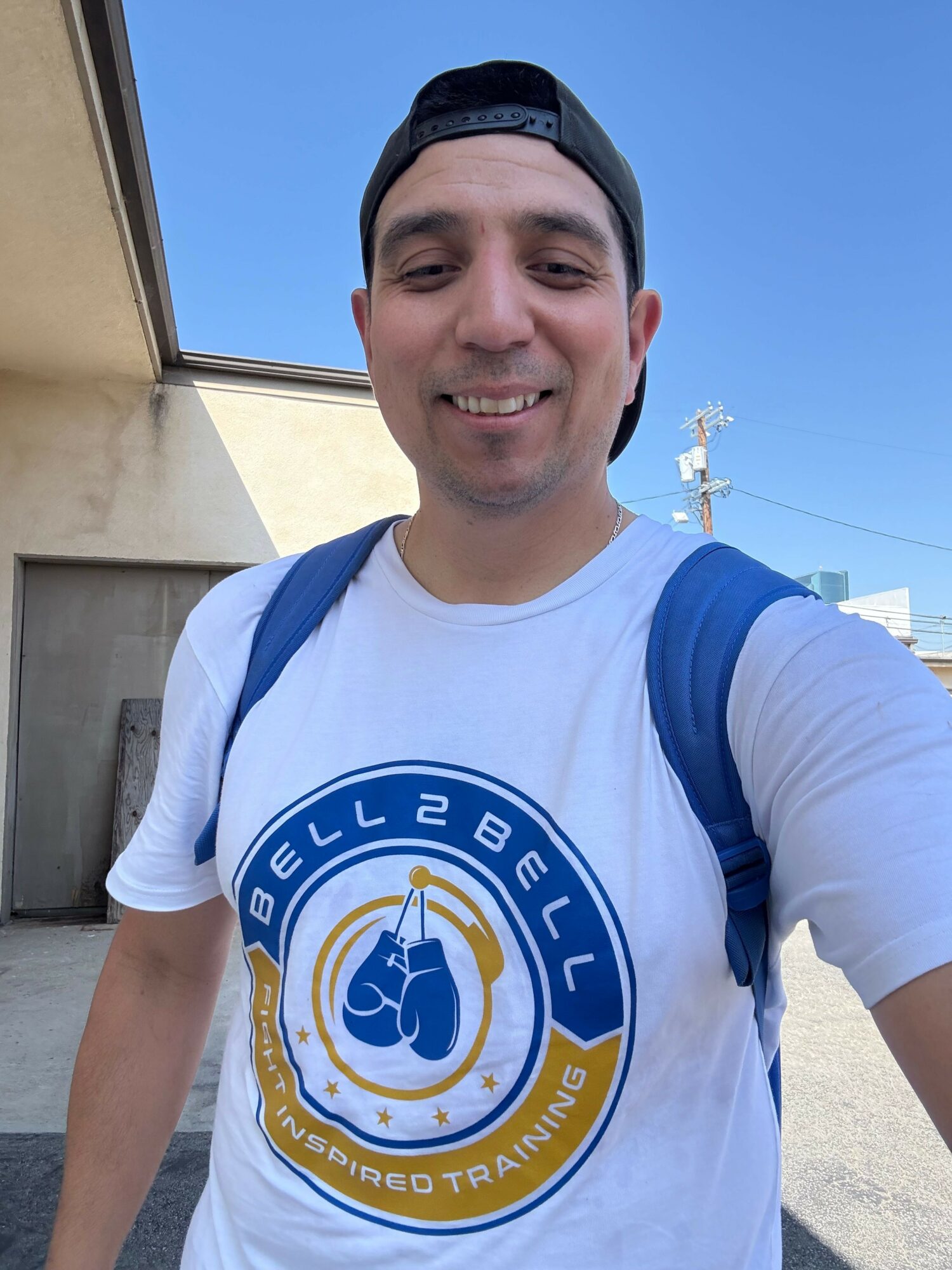 Person smiling outdoors wearing a white t-shirt with a circular logo, blue backpack, and a black cap, with a clear blue sky.