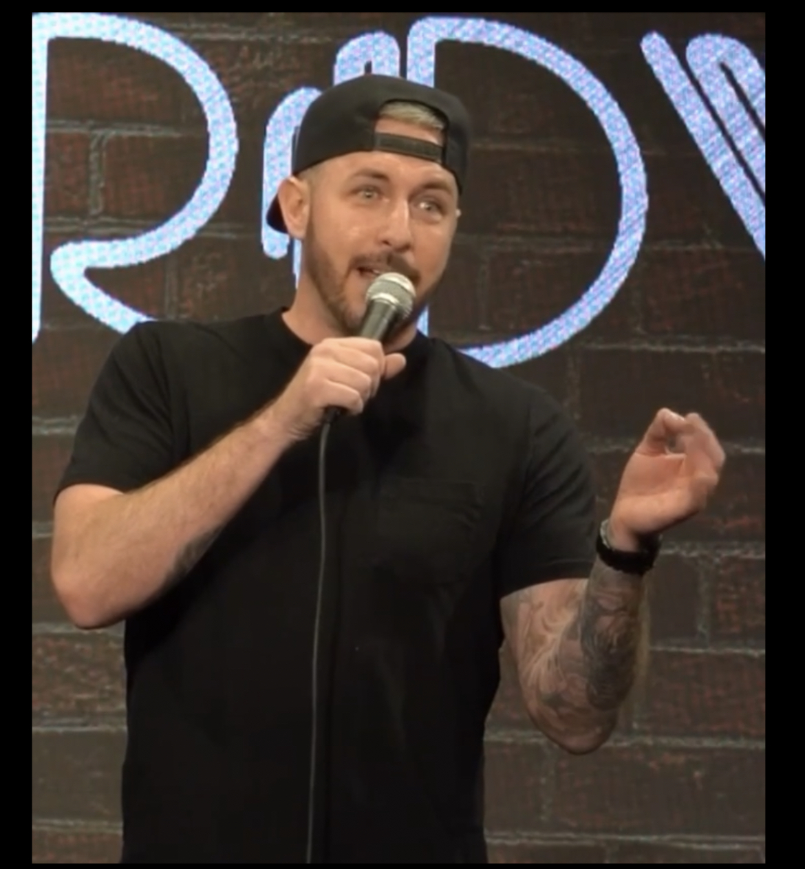 Man with a beard, wearing a black t-shirt and a backwards cap, holding a microphone and speaking on stage.