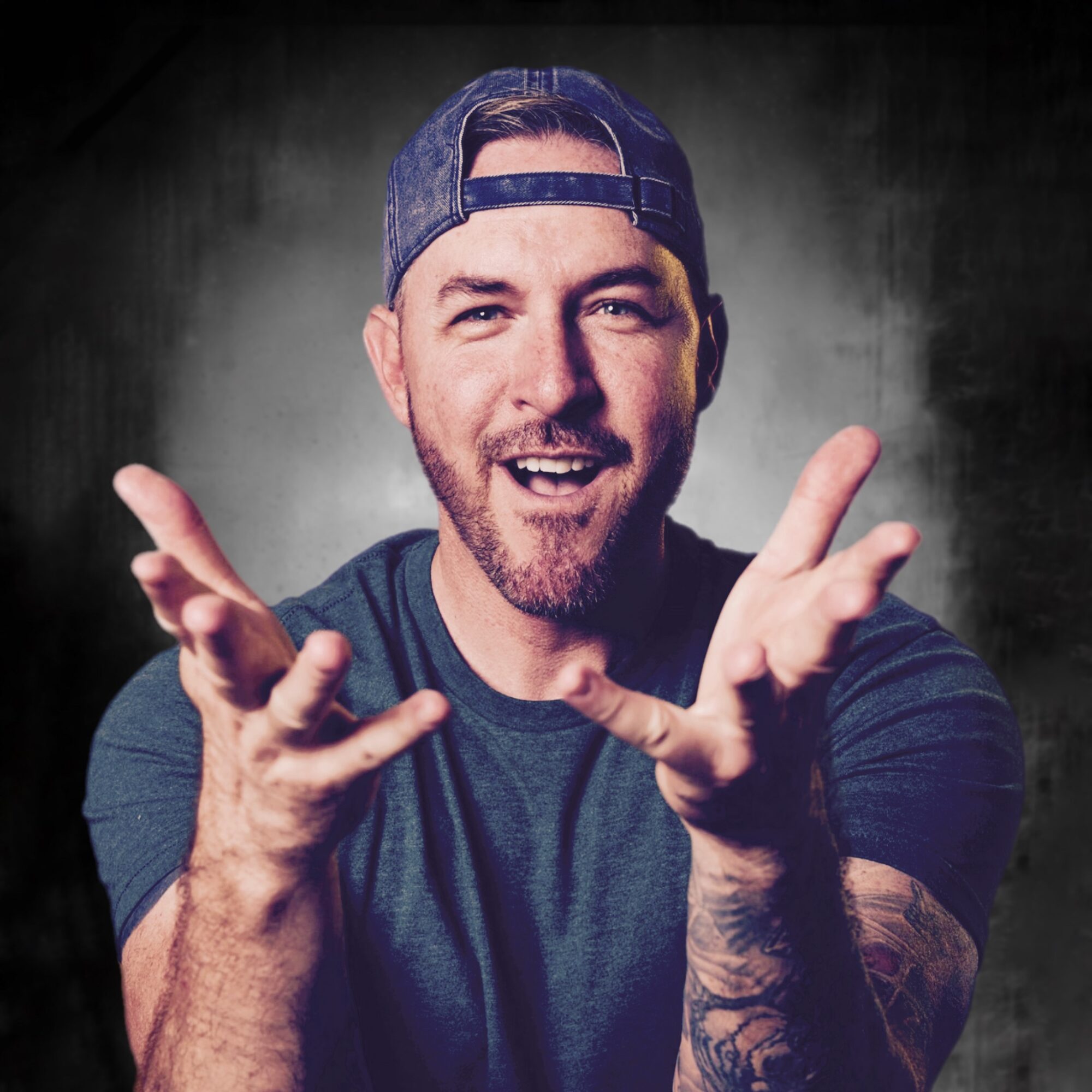 Man with a beard, wearing a backwards cap, smiling, with hands raised, against a dark background.