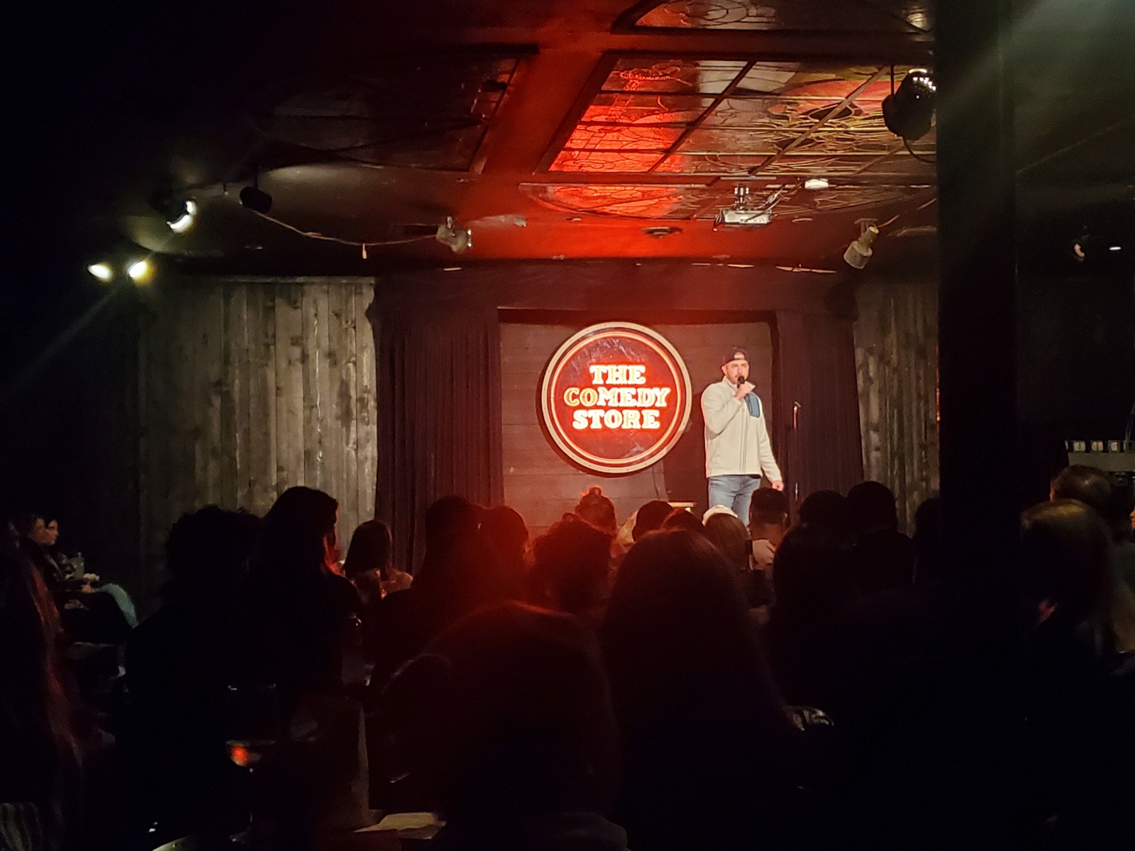 Person on stage performing in front of an audience with a circular sign reading 'The Comedy Store' behind them.