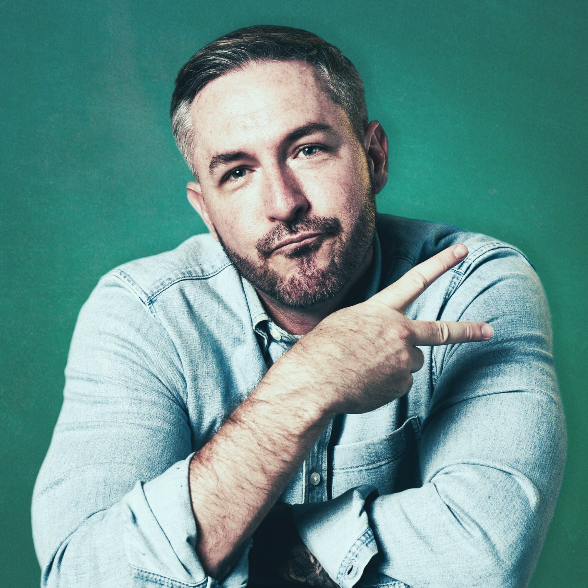 Man with short dark hair and beard, wearing a light denim shirt, pointing to the right, against a green background.