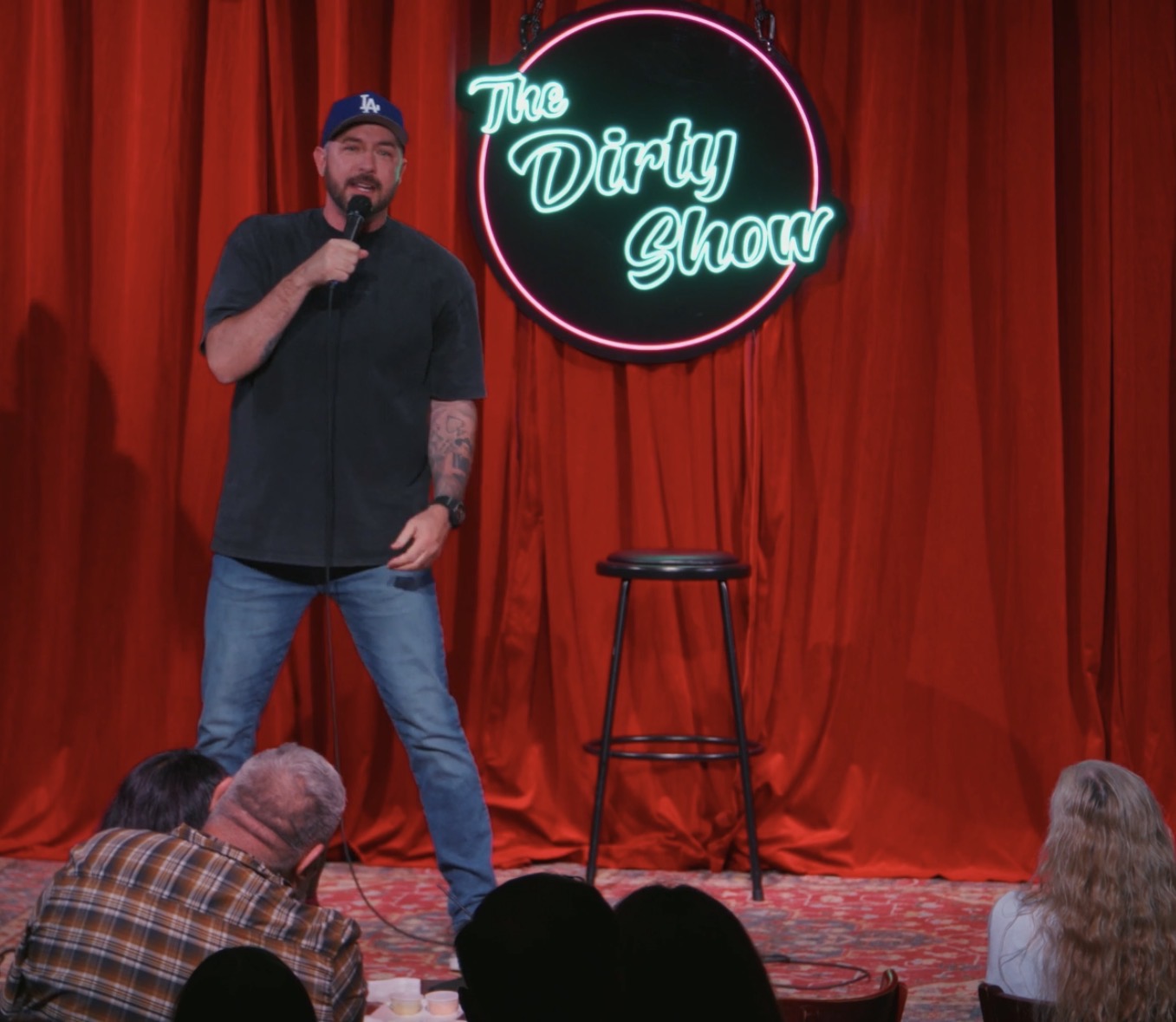 Male comedian on stage with red curtains, neon sign reading 'The Dirty Show,' and an empty stool, audience visible.