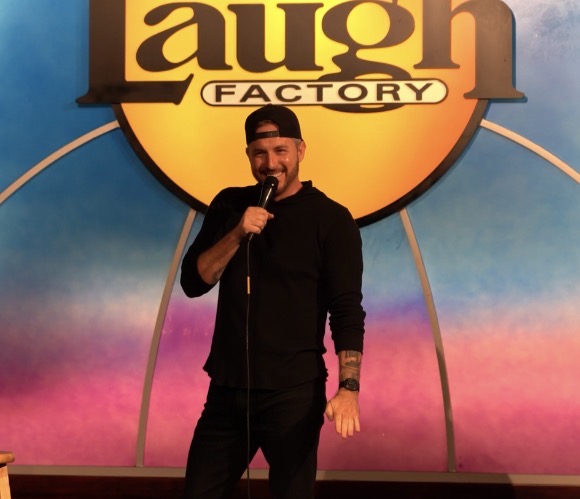 Person standing on stage holding a microphone, smiling, in front of a colorful background and a large sign reading 'laugh FACTORY'.