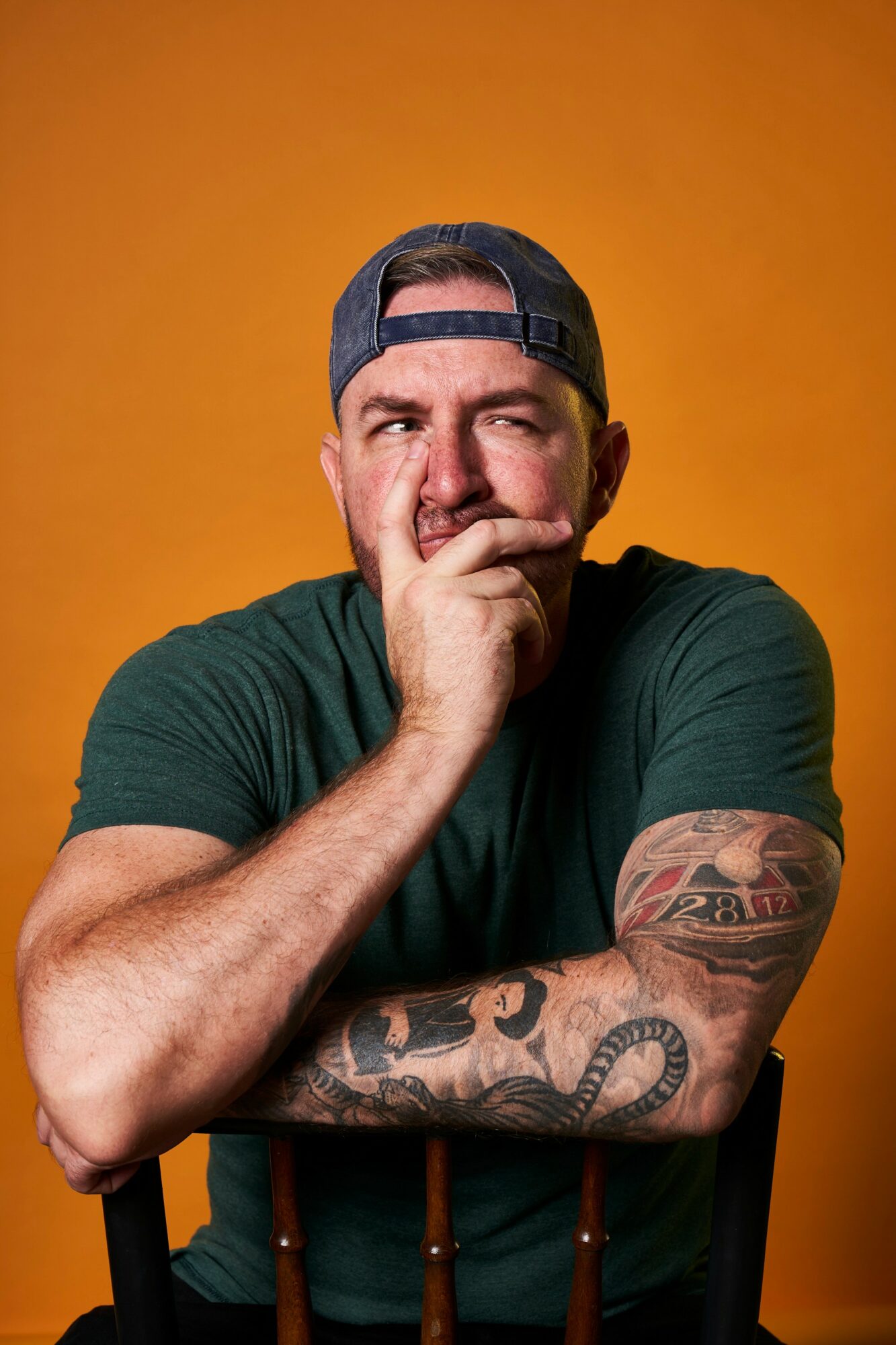 Man with a backwards cap, green shirt, and tattoos on arm, holding chin, against orange background.