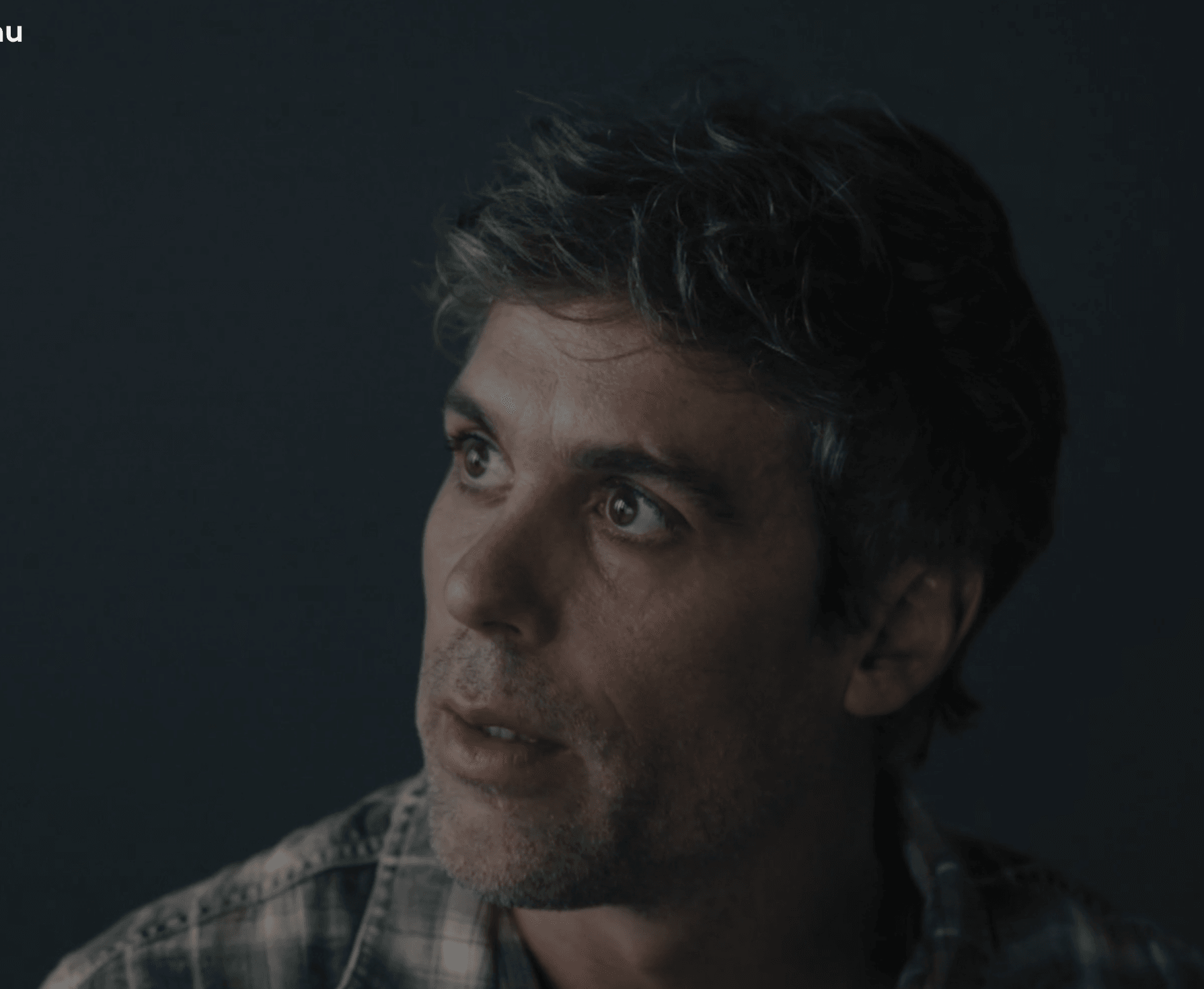 Man with dark hair and stubble looking to the side, wearing a plaid shirt, dark background