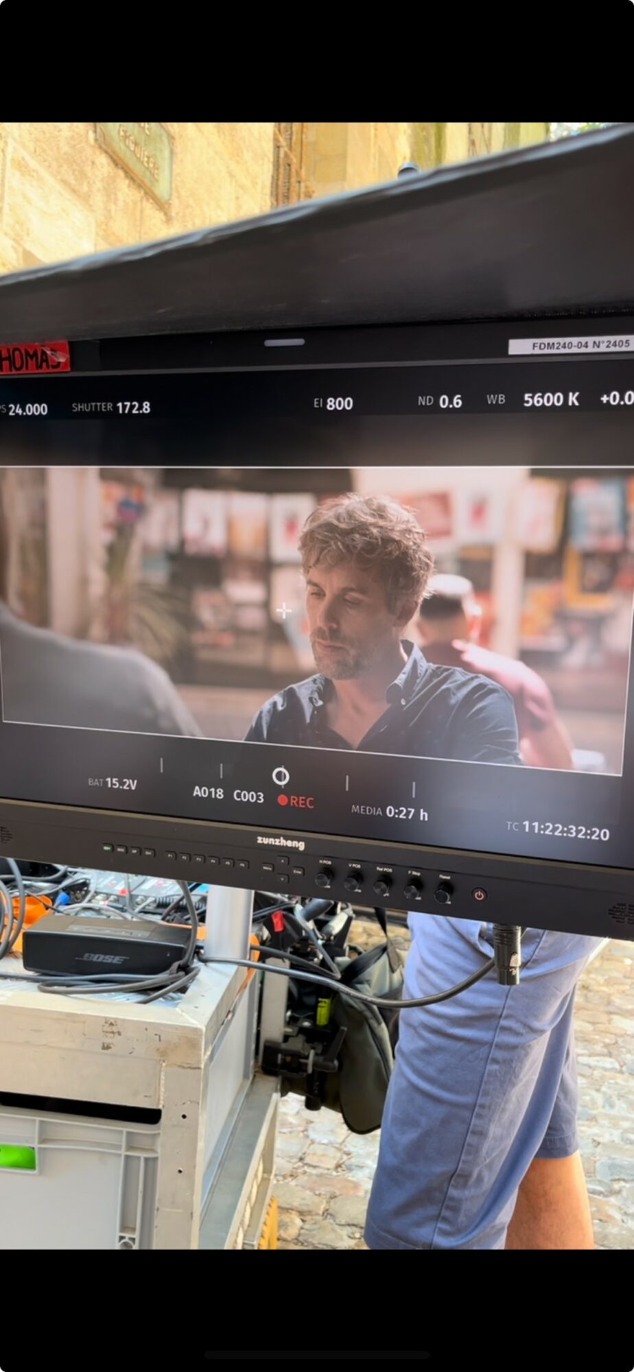 Person standing outdoors behind a camera monitor, with a busy street scene in the background.