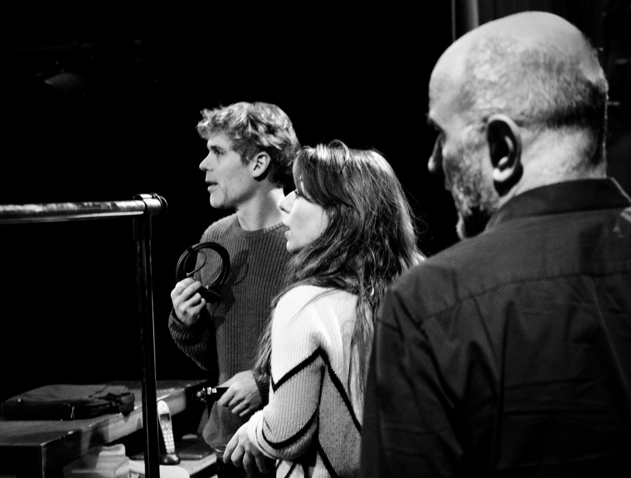 Three people in a recording studio, one with headphones, two watching, black and white image.