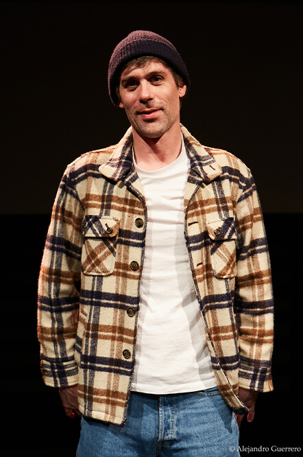 Man wearing a beanie, plaid jacket, white t-shirt, and jeans, standing against a dark background.