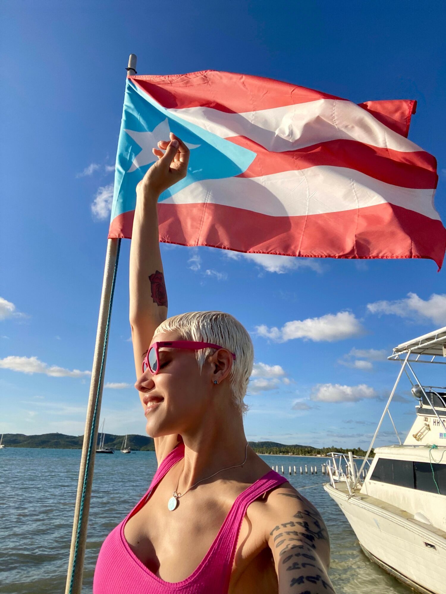 Woman with short white hair and sunglasses holding a flag with red, white, and blue stripes near a boat on water.