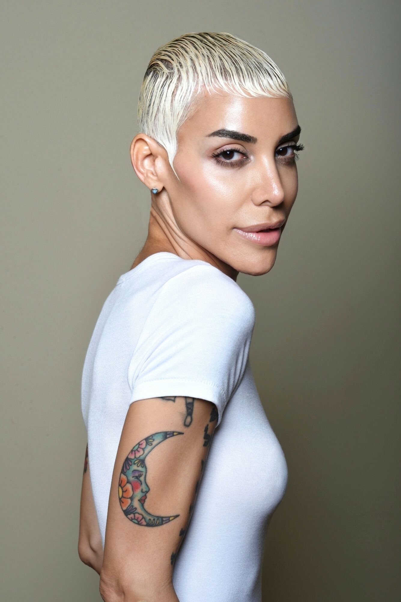 Woman with short platinum blonde hair, wearing a white shirt, showing a colorful tattoo on her upper arm, facing slightly right.