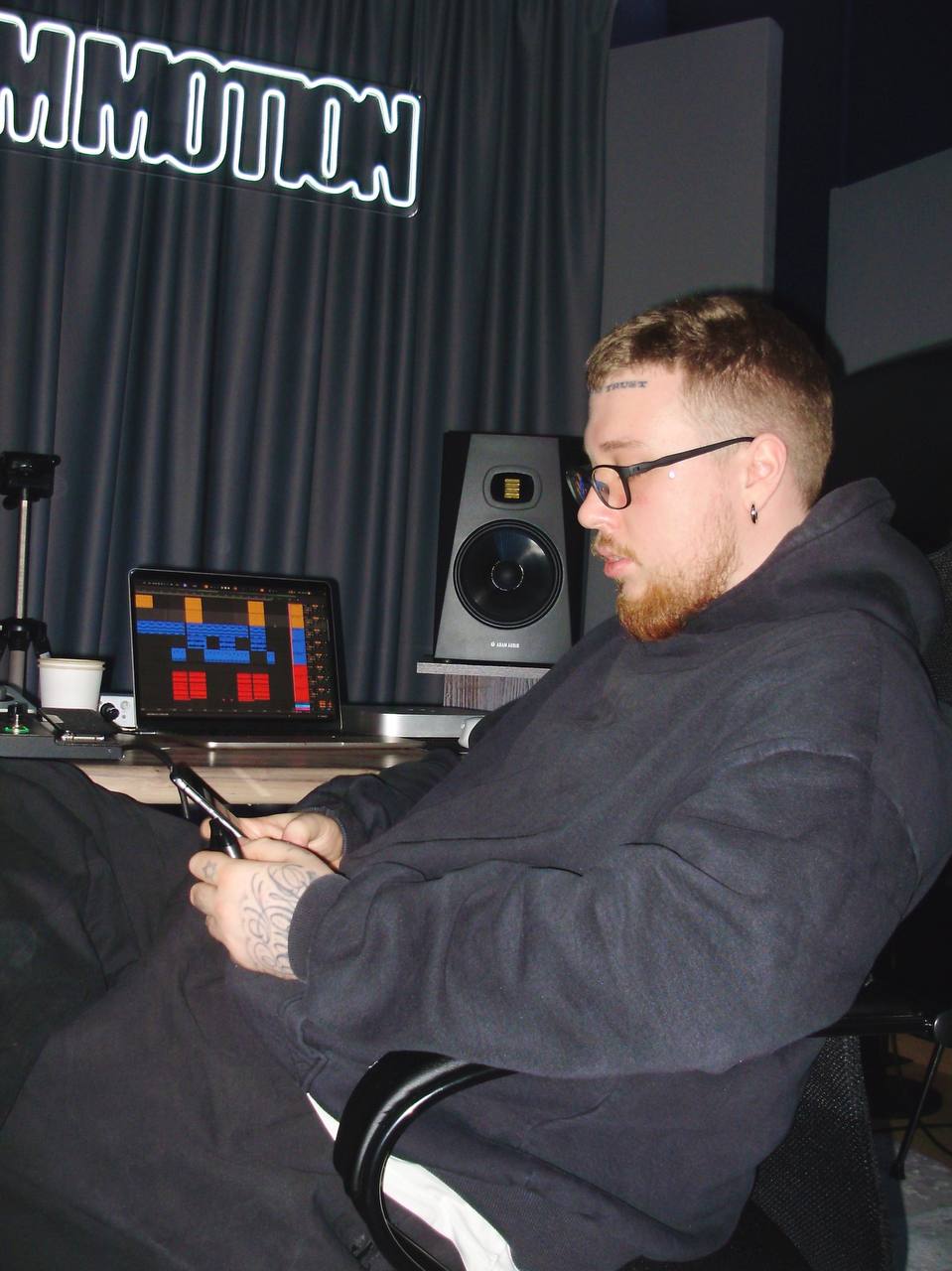 Man with glasses and a beard sitting in a music studio, looking at his phone, with audio equipment around him.