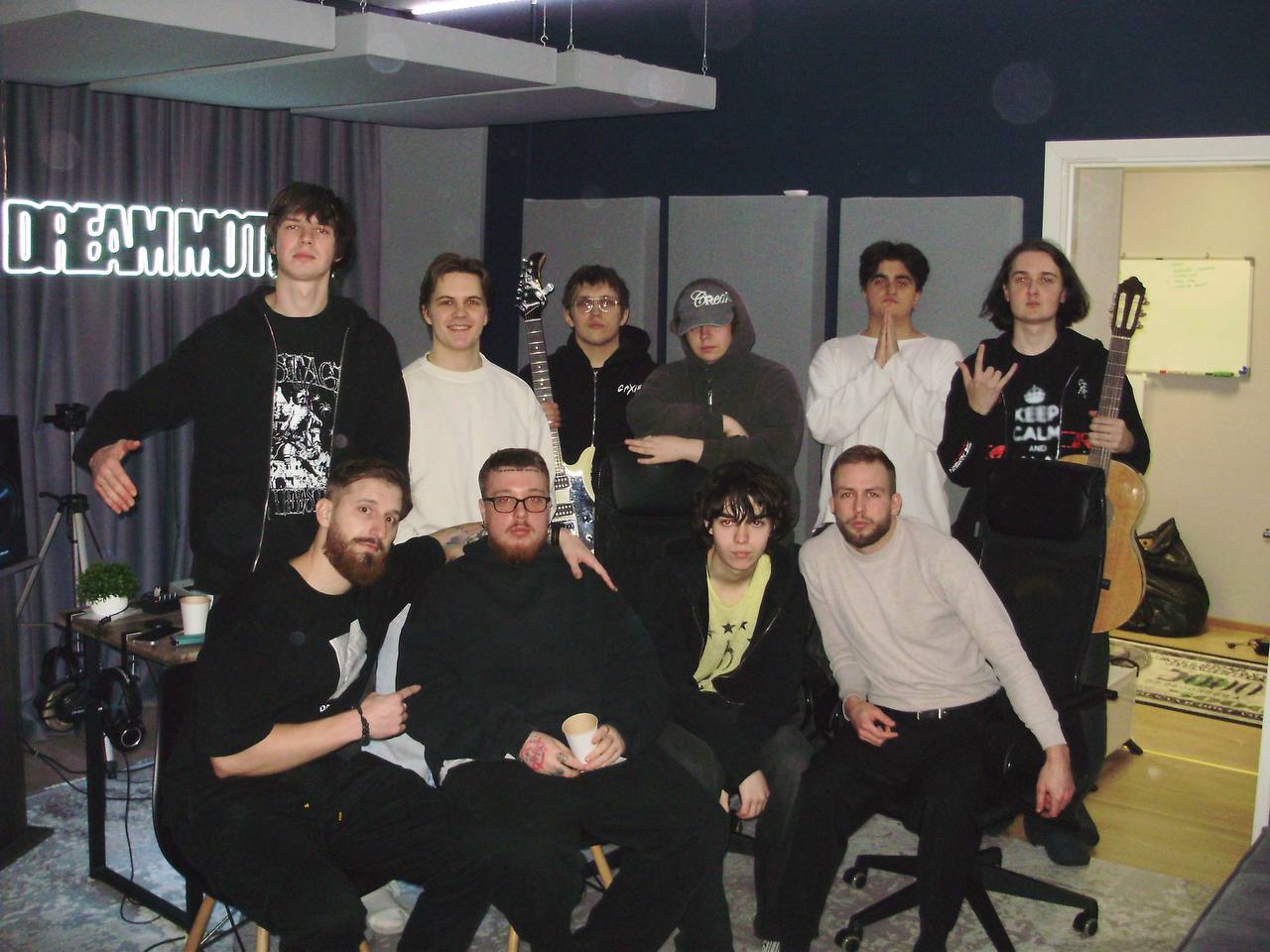 Group of ten people in a room with a dark curtain and a neon sign, some sitting and some standing, casual pose.