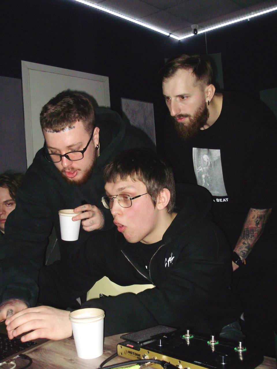 Three men gathered around a table with drinks, one holding a cup, in a dark room with neon lighting.