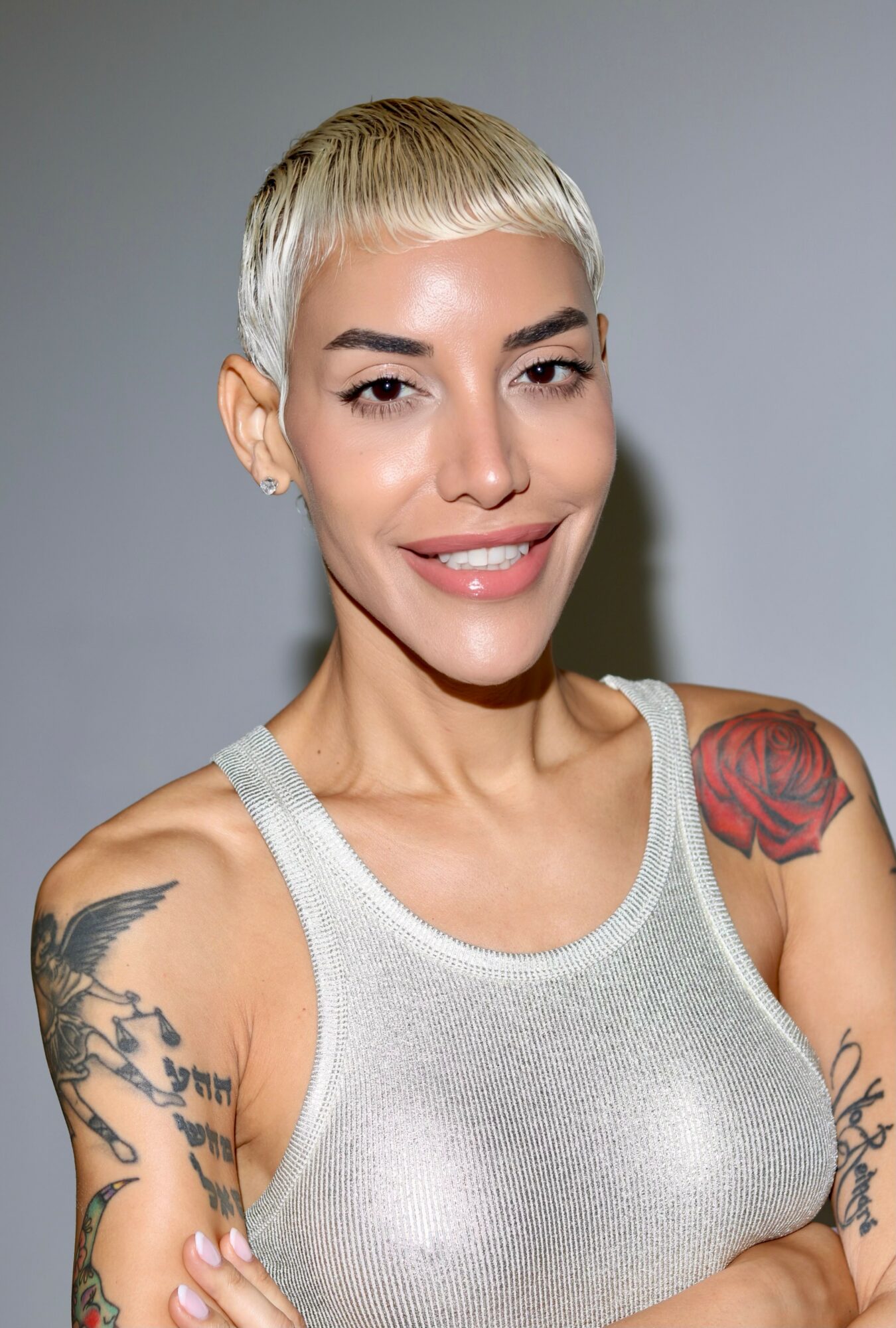 Woman with short platinum blonde hair, smiling, wearing a gray tank top, with tattoos on her arms and shoulder.