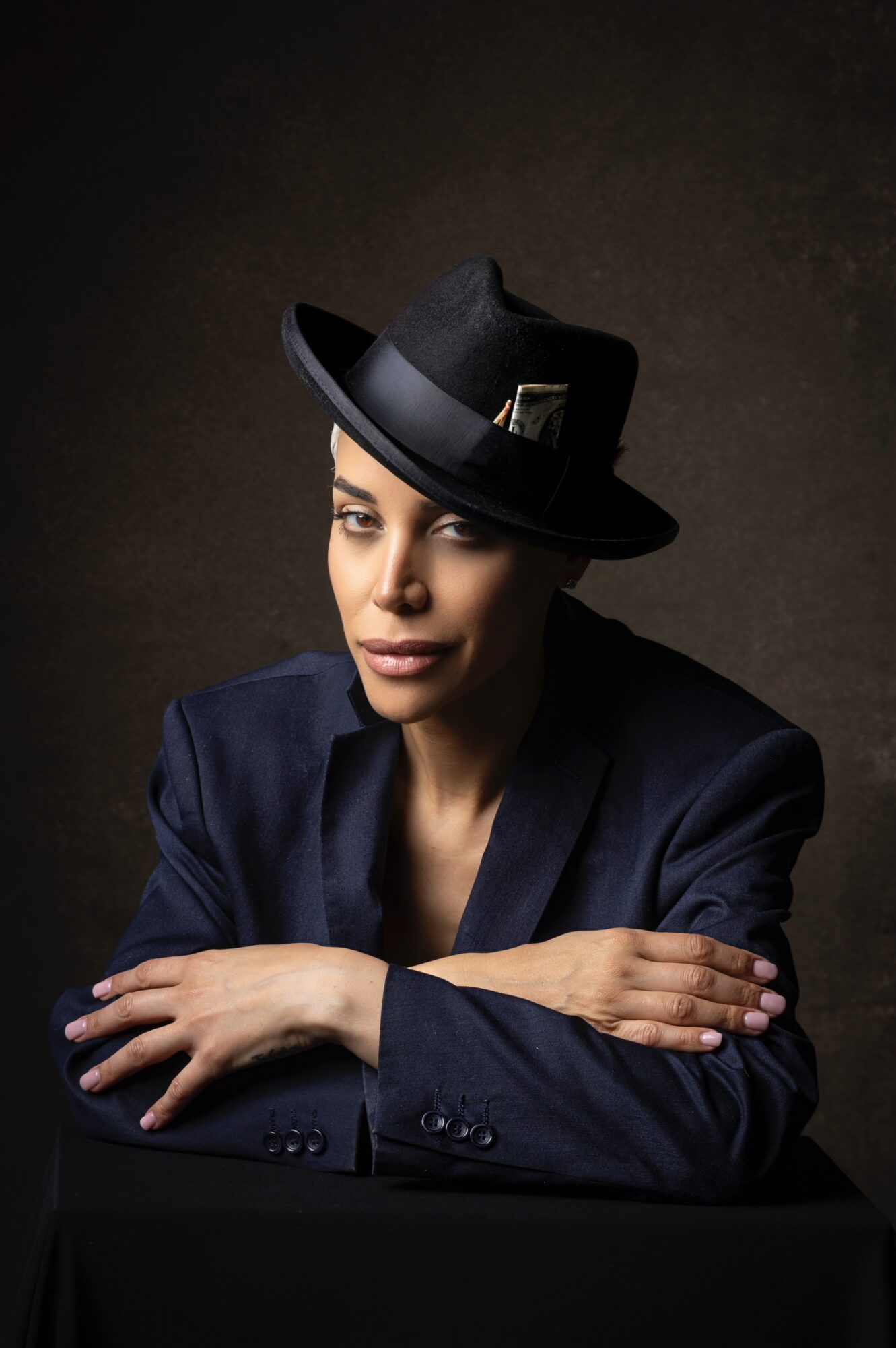 Woman wearing a dark hat with a gold buckle, dark blazer, resting her arms on a surface, against a dark background.