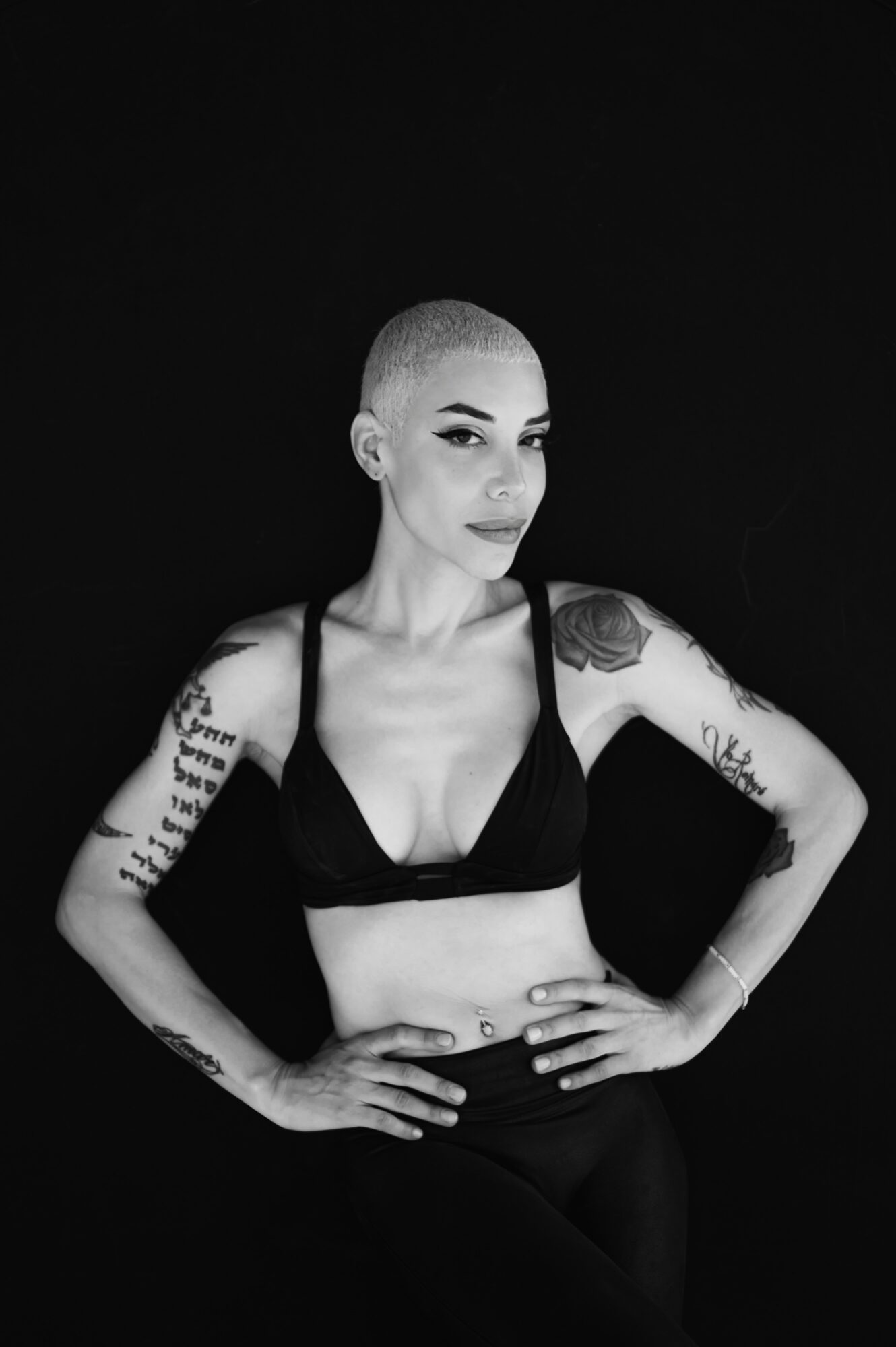 Woman with a shaved head and tattoos, wearing a black top, standing with hands on hips against a black background.