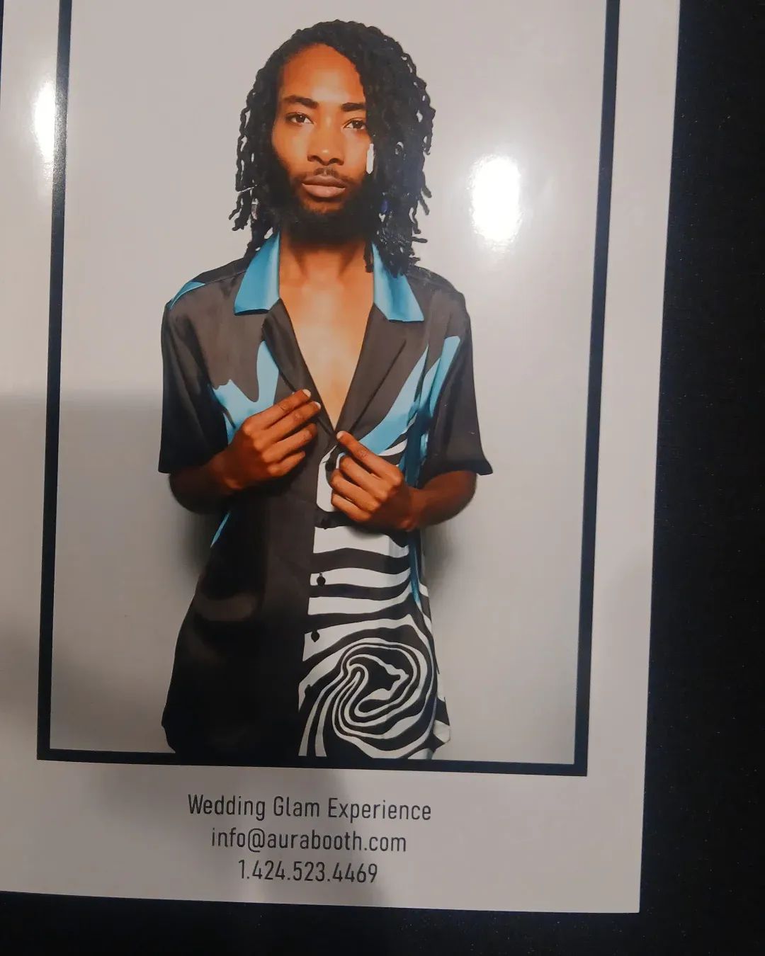 Person with shoulder-length curly hair wearing a patterned shirt, holding the collar, standing against a plain background.