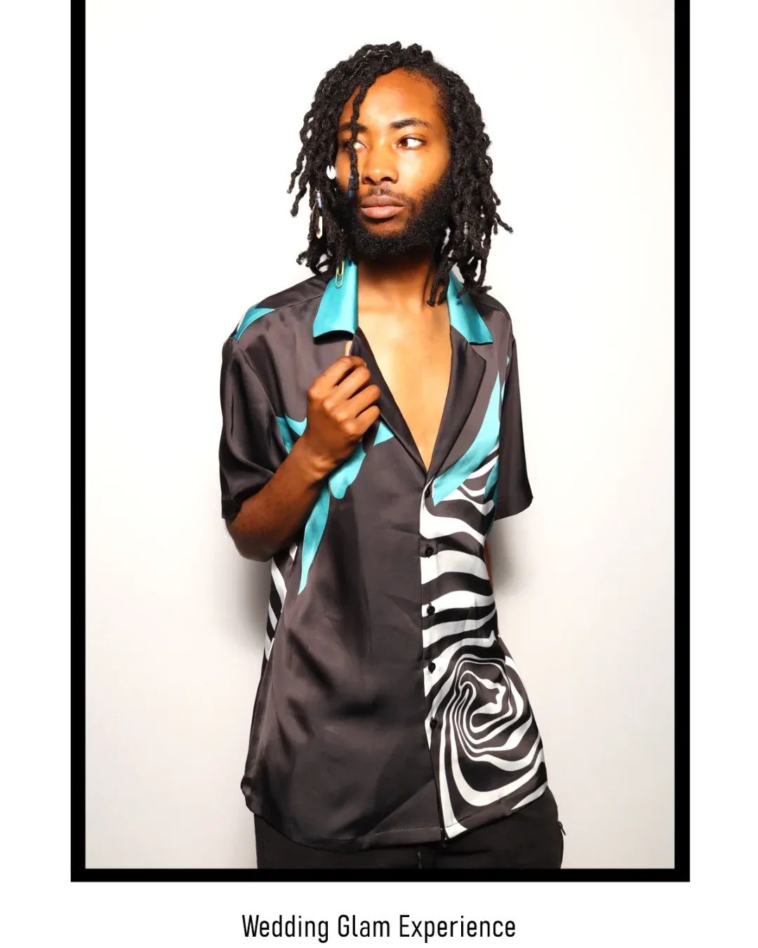 Man with dreadlocks wearing a black and teal patterned shirt, standing against a white background.