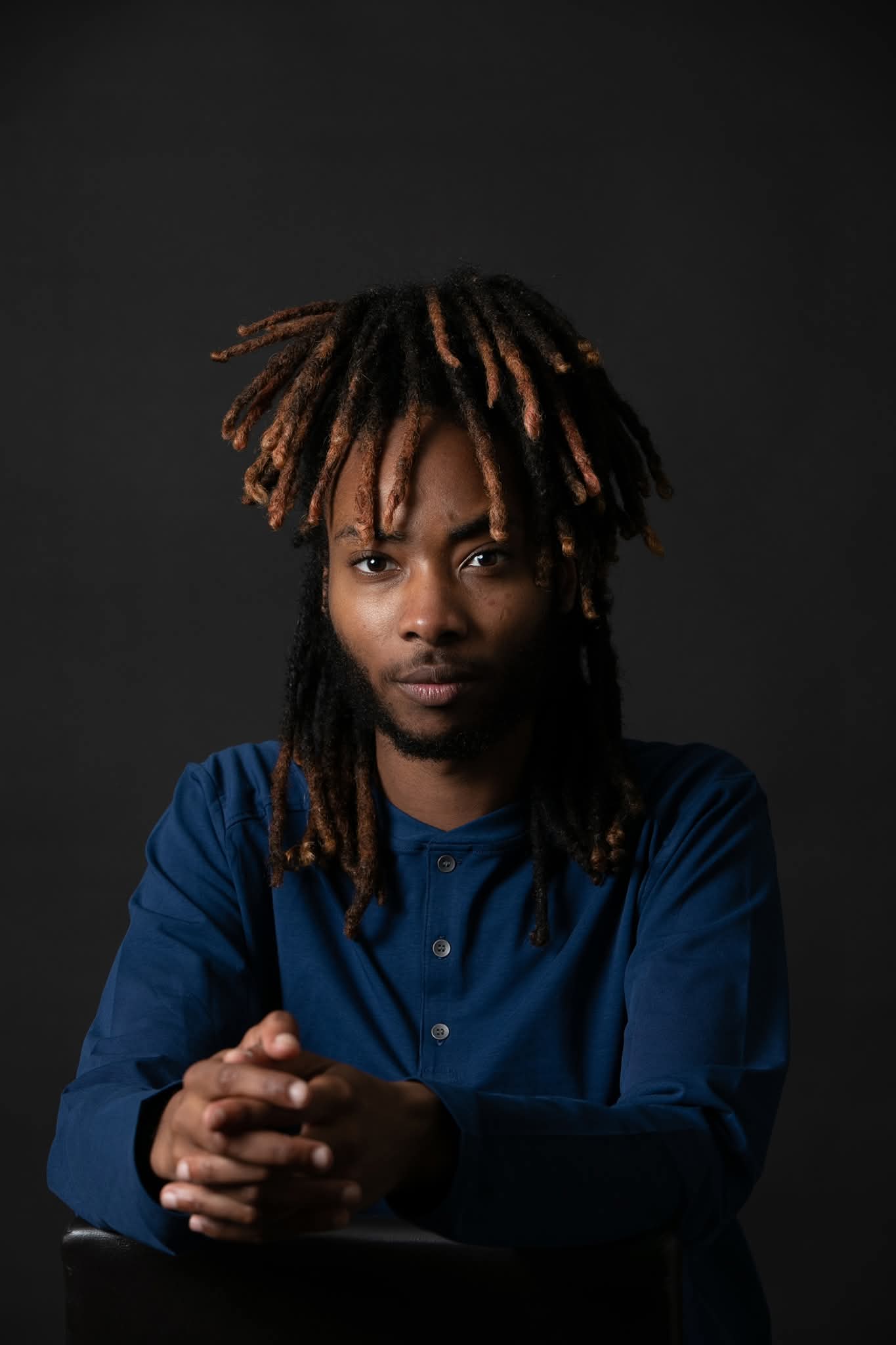Person with dreadlocks wearing a blue shirt, looking directly at the camera, hands clasped, dark background.