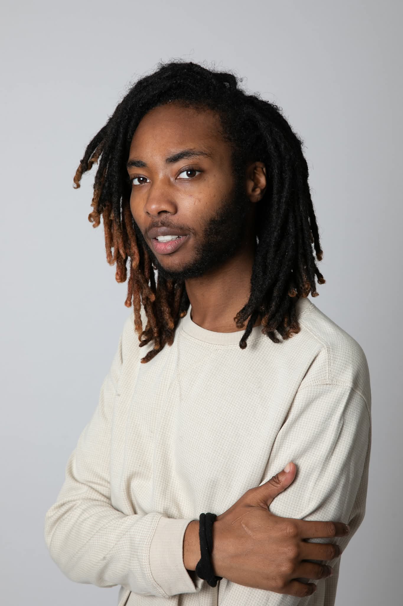Young man with dreadlocks wearing a light-colored sweatshirt, standing against a plain background.