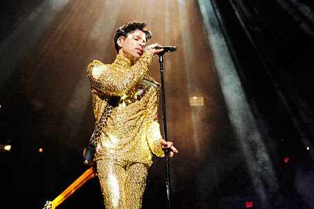 Person singing into a microphone on stage, wearing a gold sequined suit, with stage lights shining down.