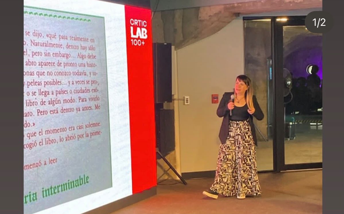 Woman standing near a large screen displaying text, in a room with glass doors and a red sign reading 'CRITIC LAB'.